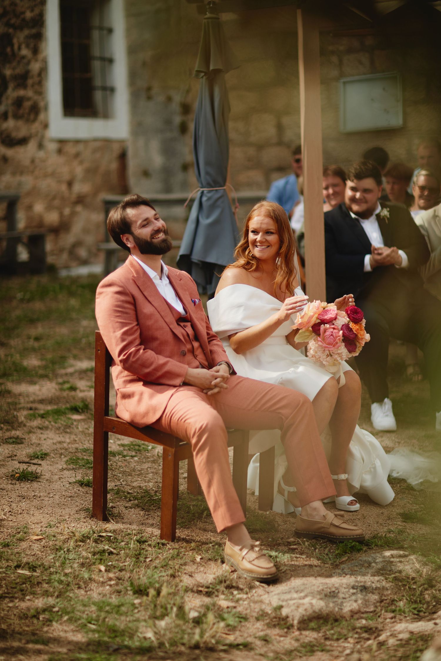 Joyful bride holding pink bouquet and bearded groom in salmon suit seated together at outdoor wedding.