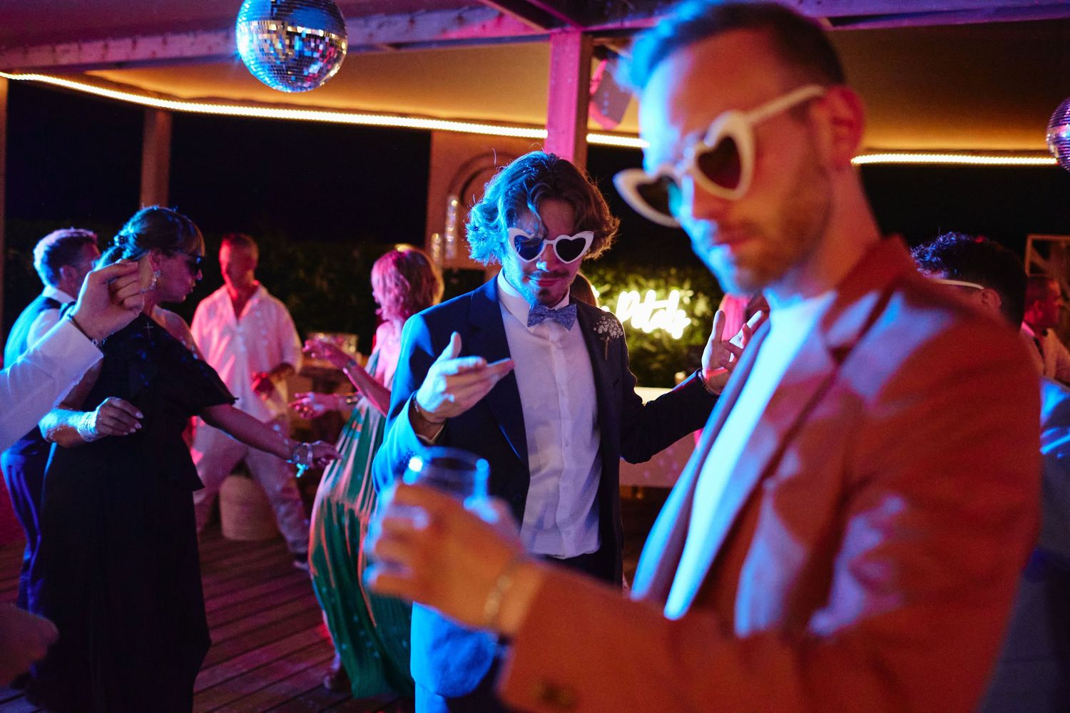 Men dancing at a lively wedding reception party under disco ball lights with colorful neon lighting.