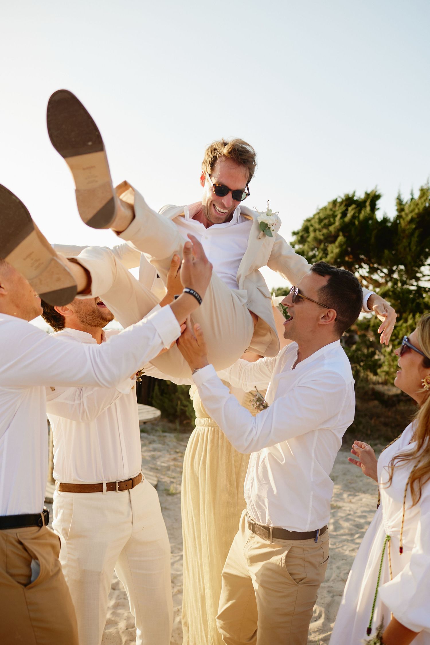 Groomsmen lifting a laughing groom in a beige suit into the air on a sandy beach.