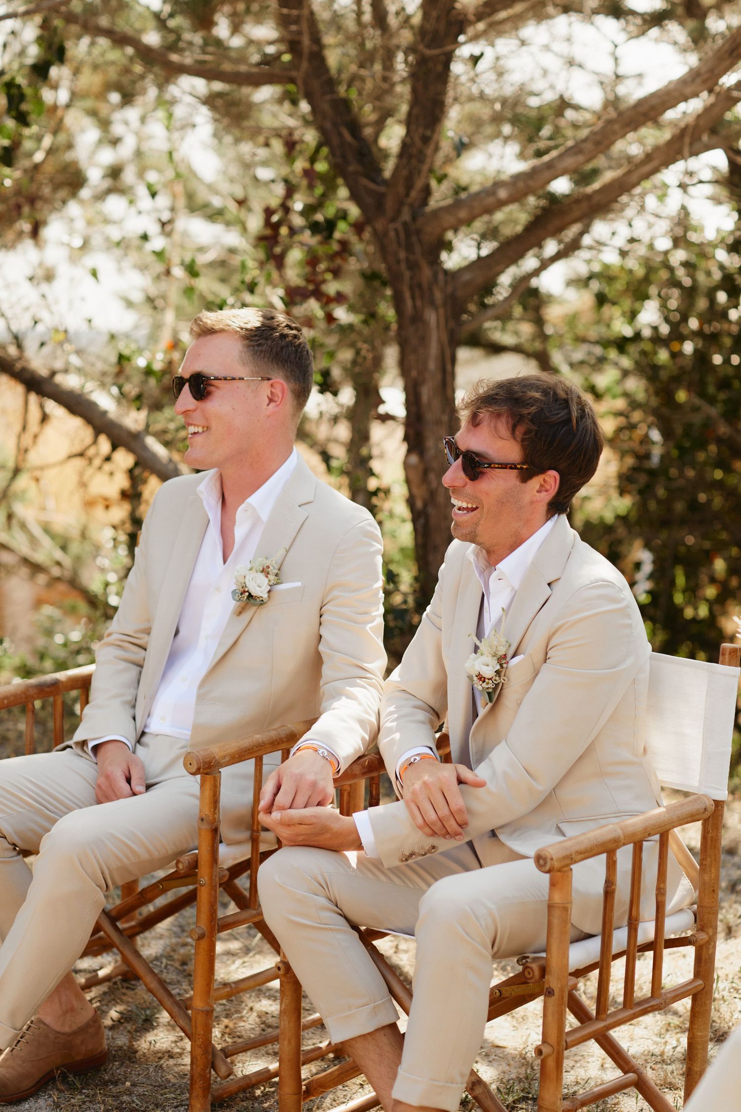 Two grooms in beige suits laughing together at outdoor woodland wedding ceremony.