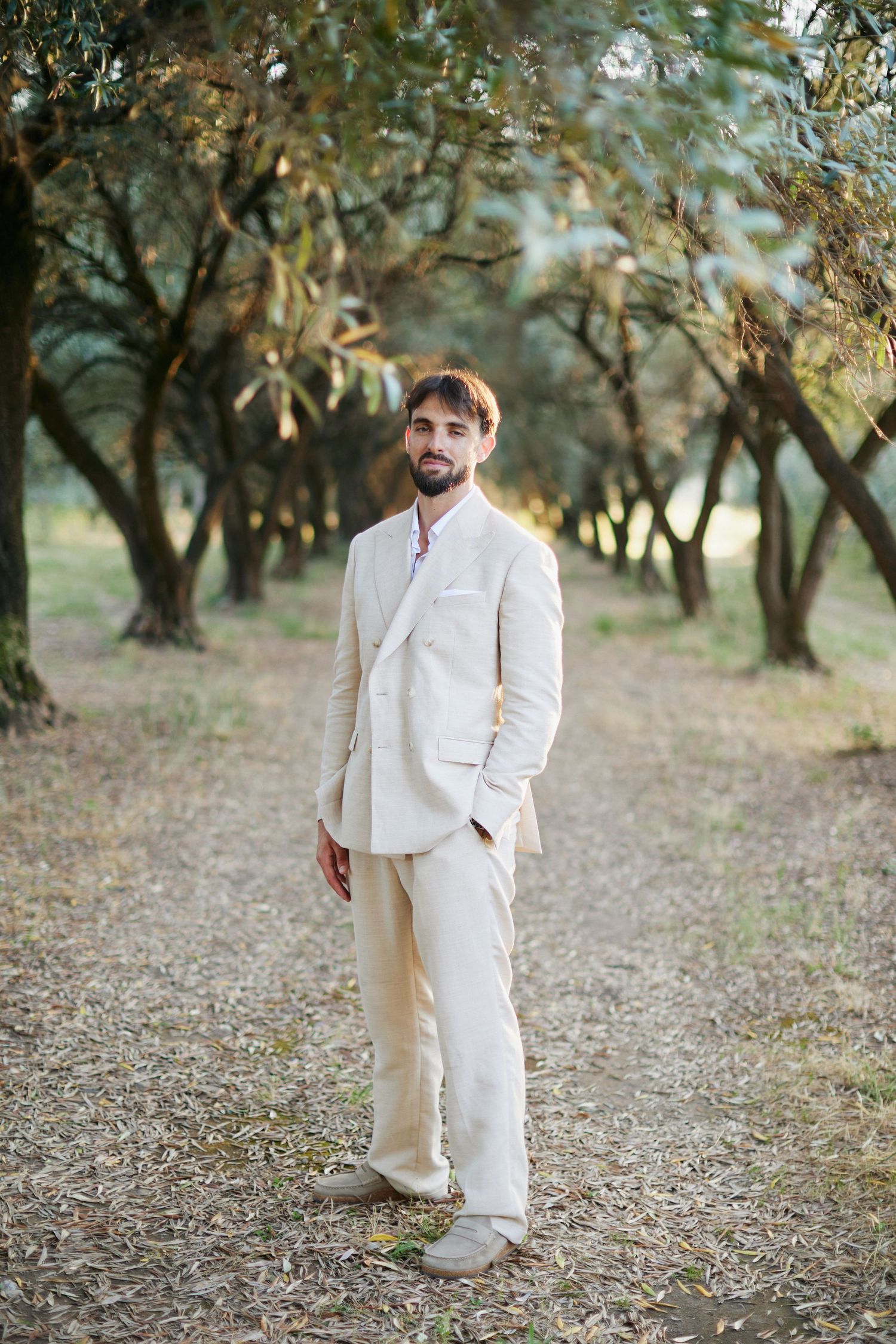 Handsome groom in beige linen suit standing confidently on a tree-lined path in a sunlit grove.