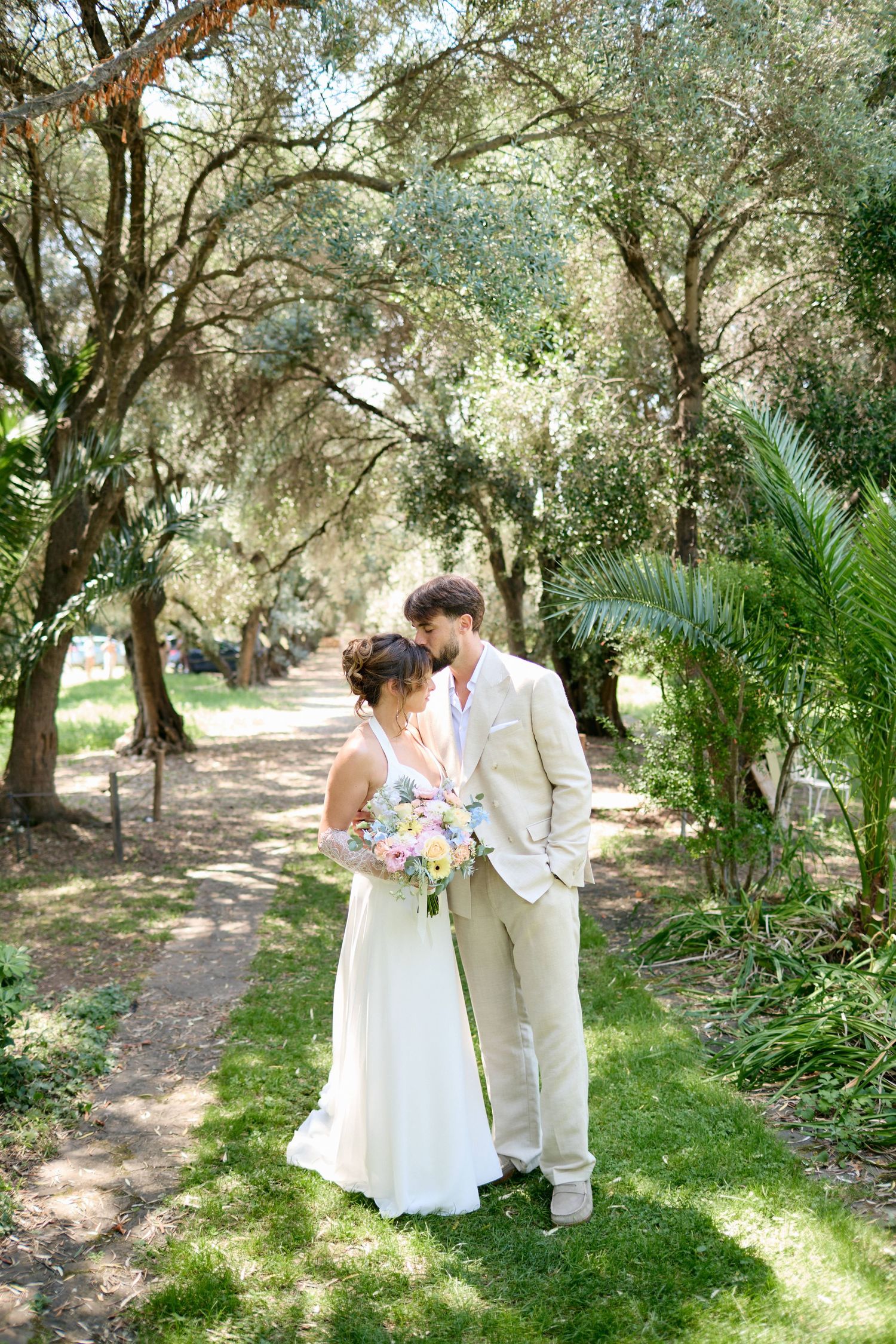 Bride and groom sharing a romantic moment on a sunlit garden path surrounded by trees.