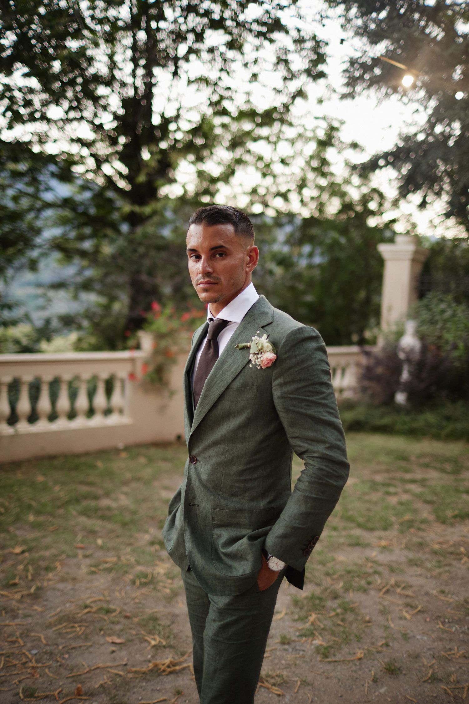 Dapper groom poses confidently in olive green linen suit with floral boutonniere in elegant garden setting.