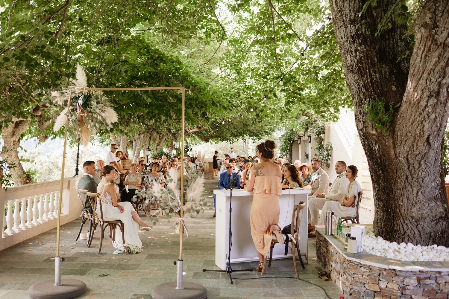 Outdoor wedding ceremony under lush green trees with guests seated at elegant venue.