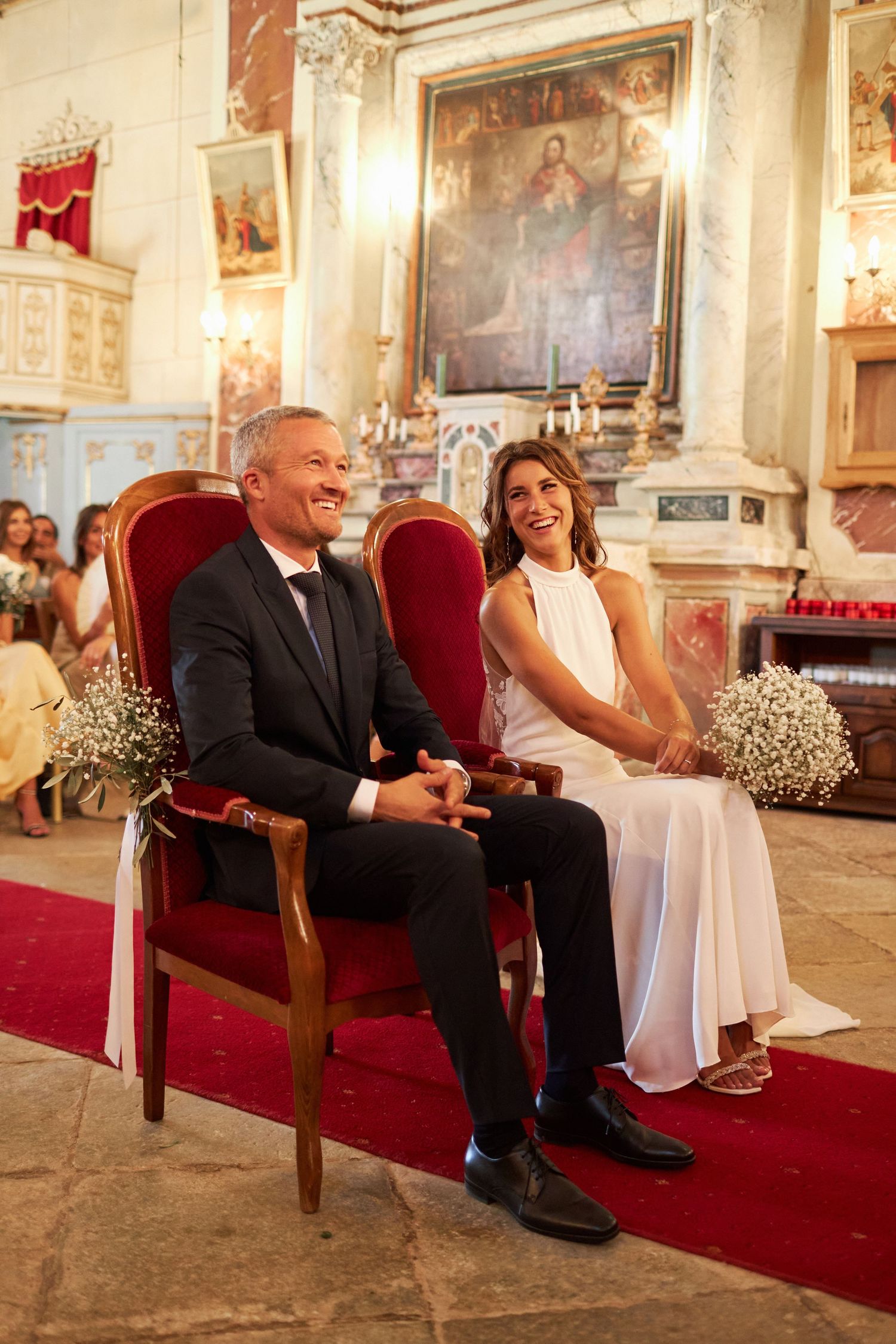 Bride and groom seated on ornate red chairs at church altar, smiling during wedding ceremony.