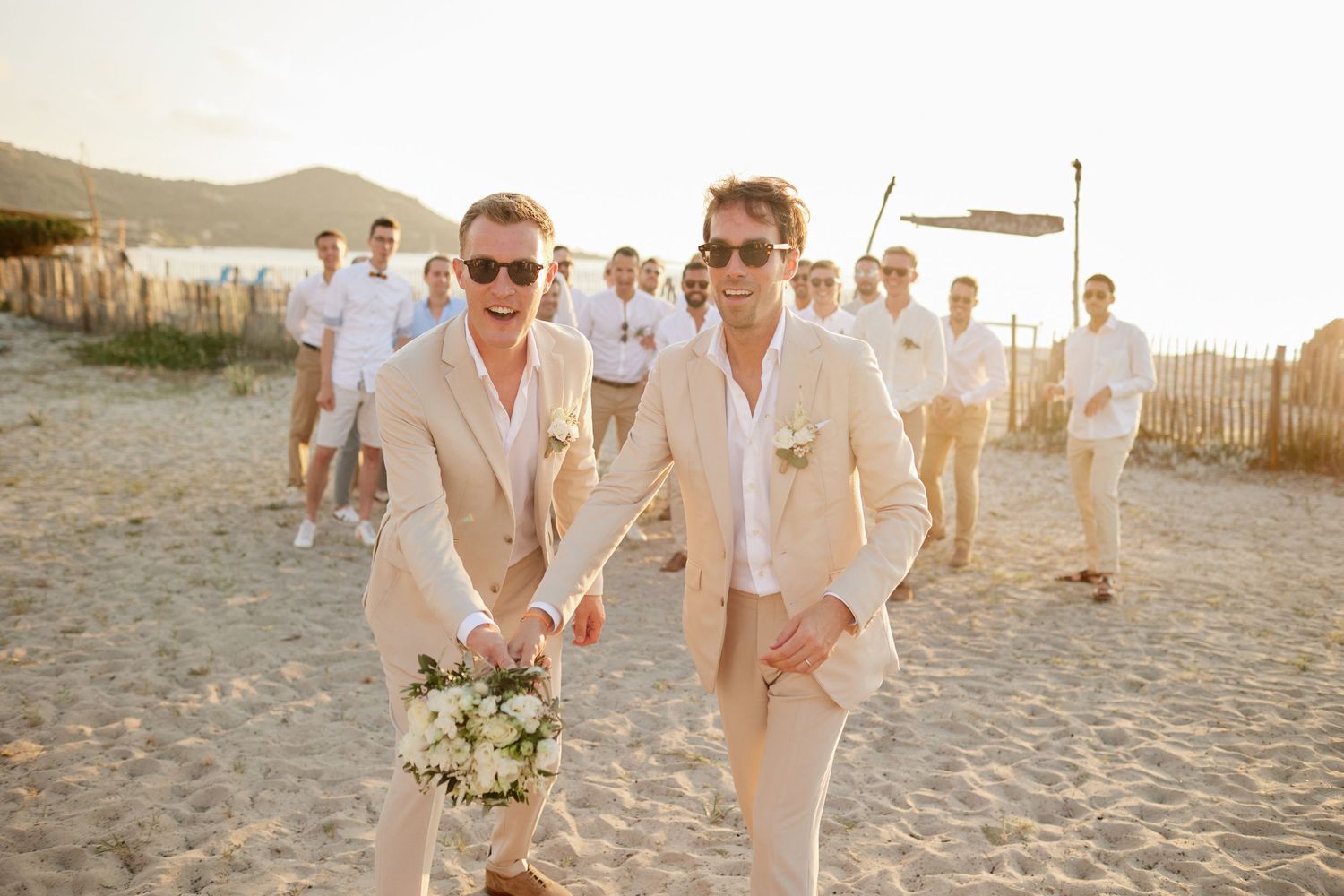 Two grooms in tan suits holding a bouquet walking on a sunny beach with wedding party behind them.