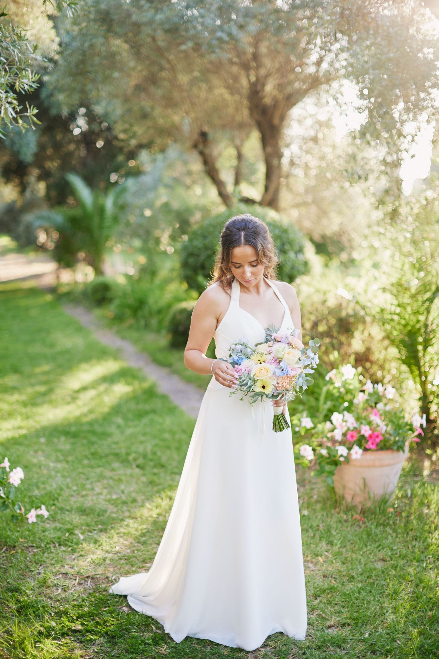 Bride in white halter gown holds pastel bouquet in sunlit garden.