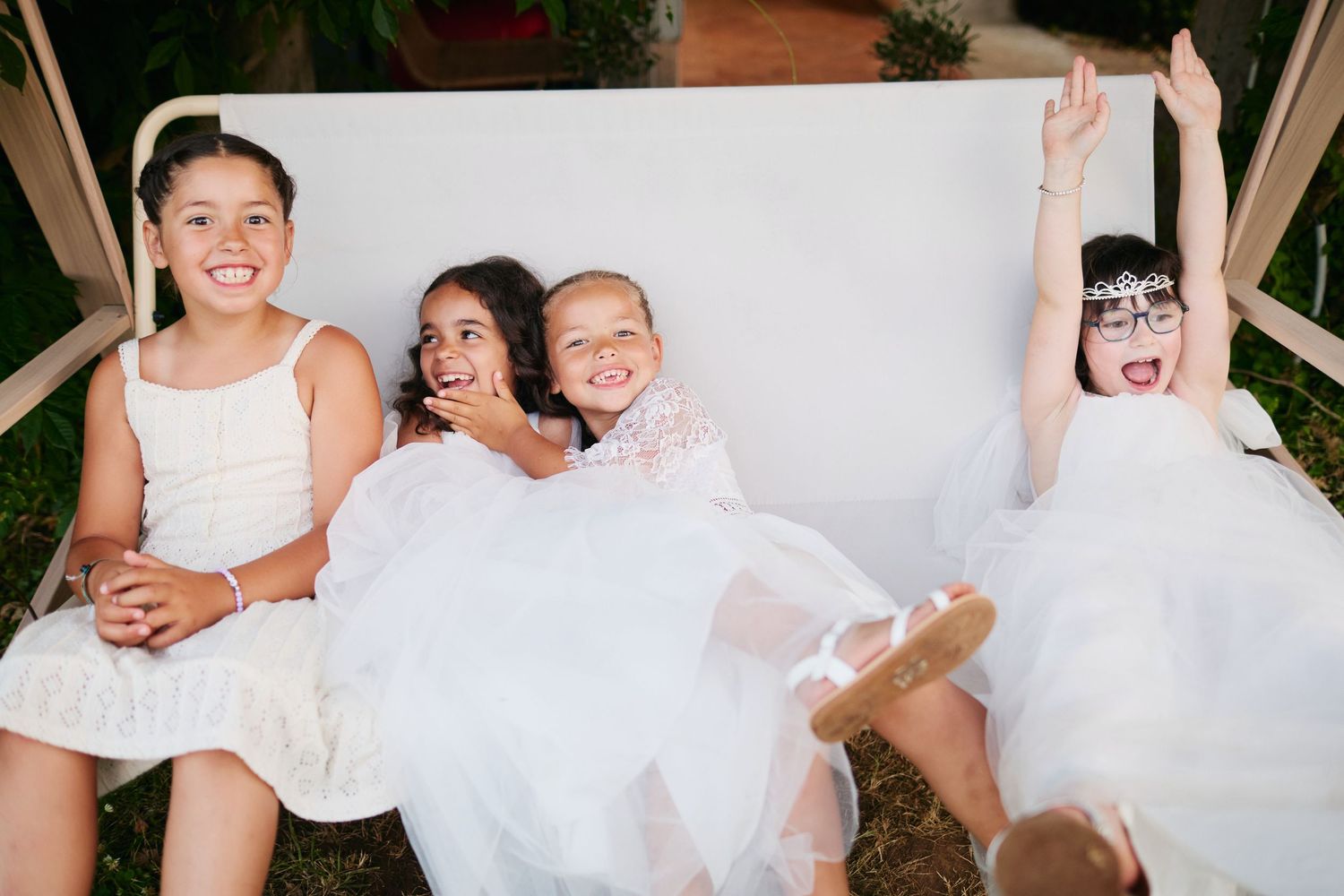 Four young flower girls in white dresses laughing and playing on a white bench at outdoor wedding.