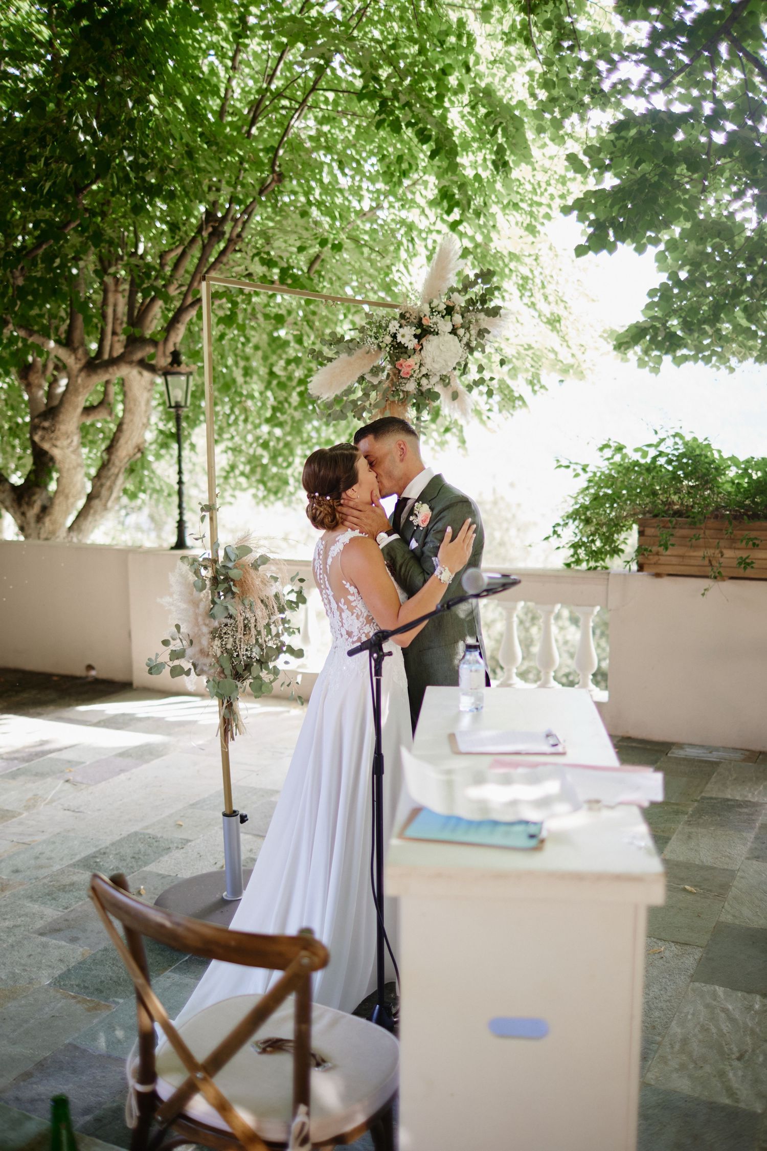 Newlyweds sharing first kiss at outdoor wedding ceremony beneath floral arch and lush green tree canopy.