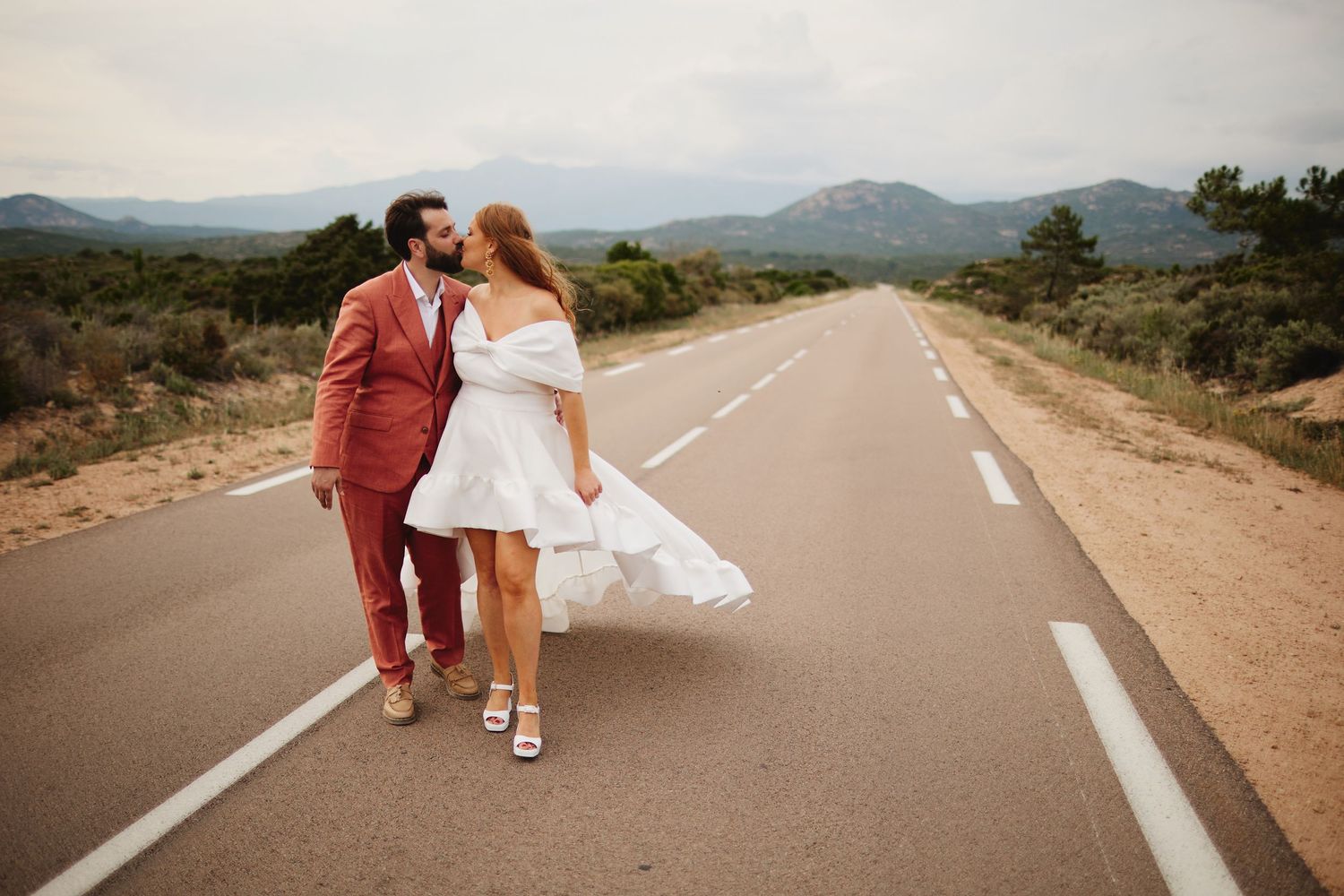 Bride and groom share a kiss while walking down a scenic empty road framed by mountains.