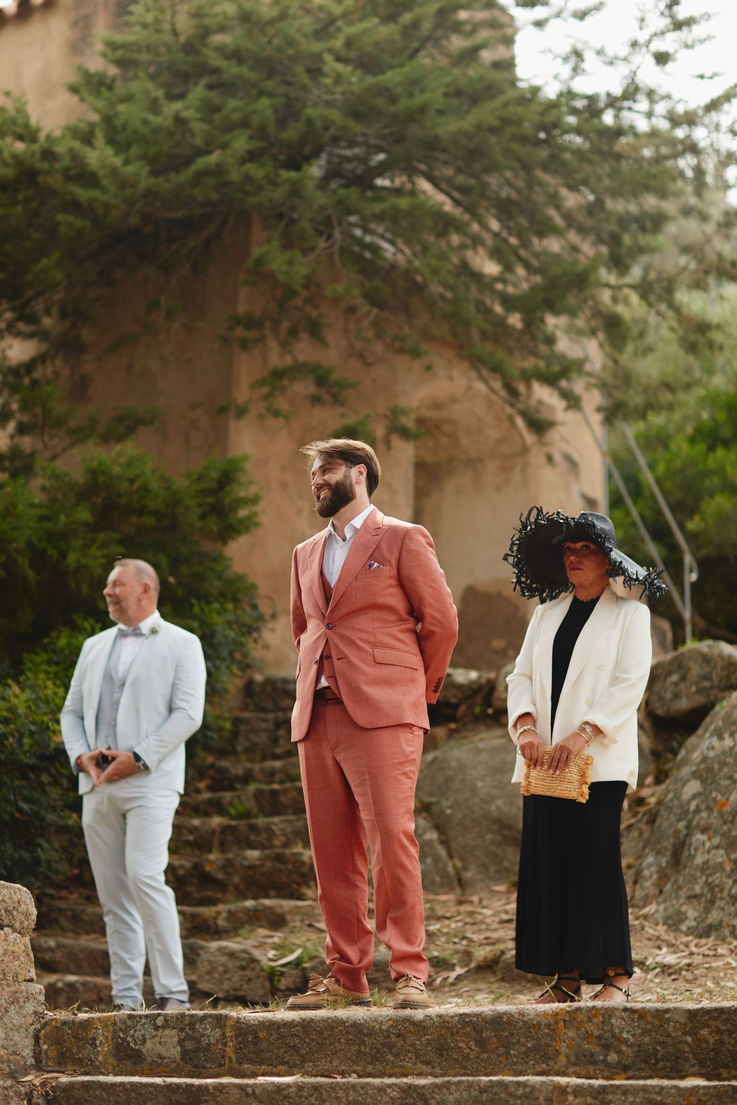 Wedding guests in coral suit and black hat stand on stone steps at rustic outdoor venue.