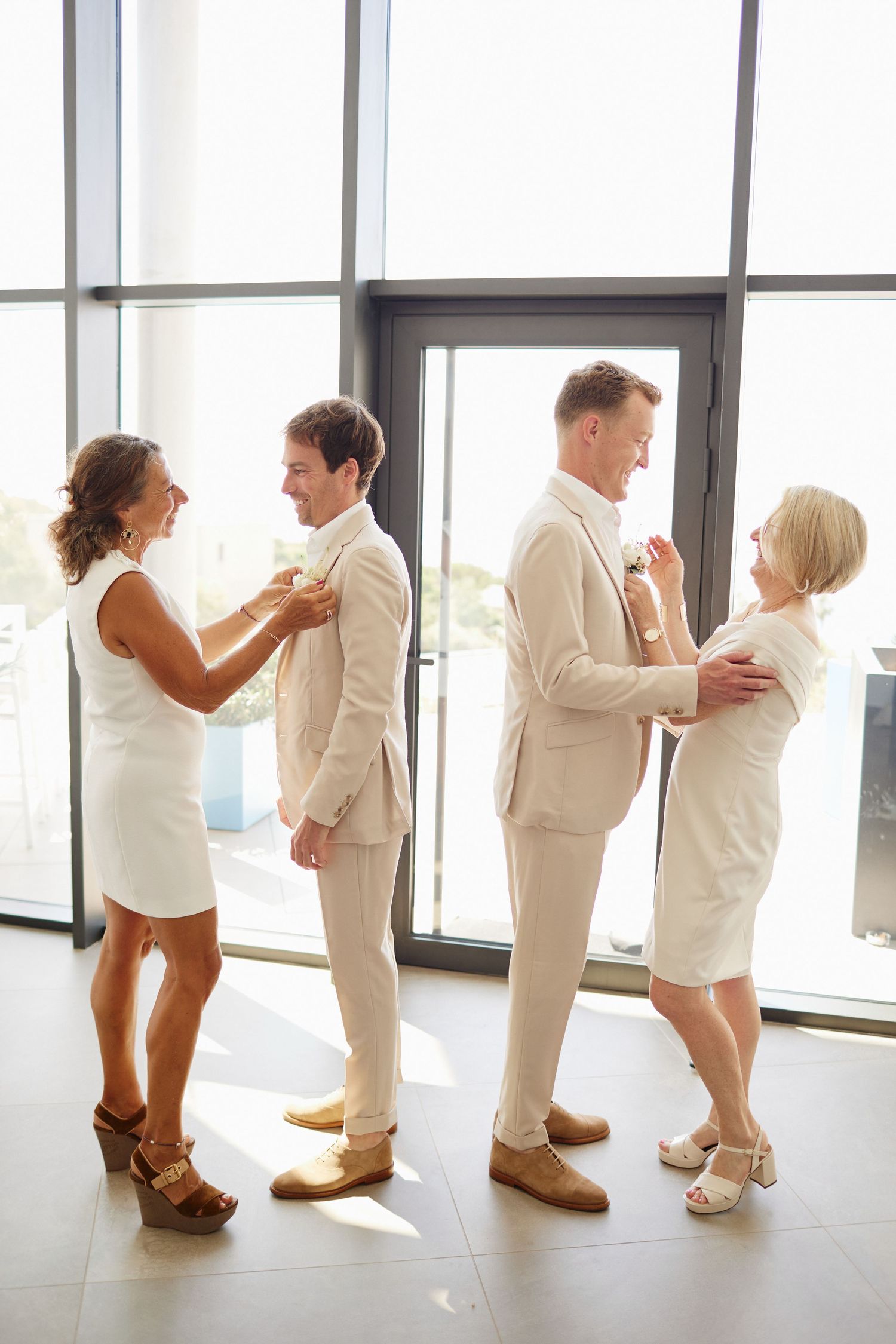 Two grooms in beige suits having boutonnieres pinned by mothers in white dresses in sunlit room.