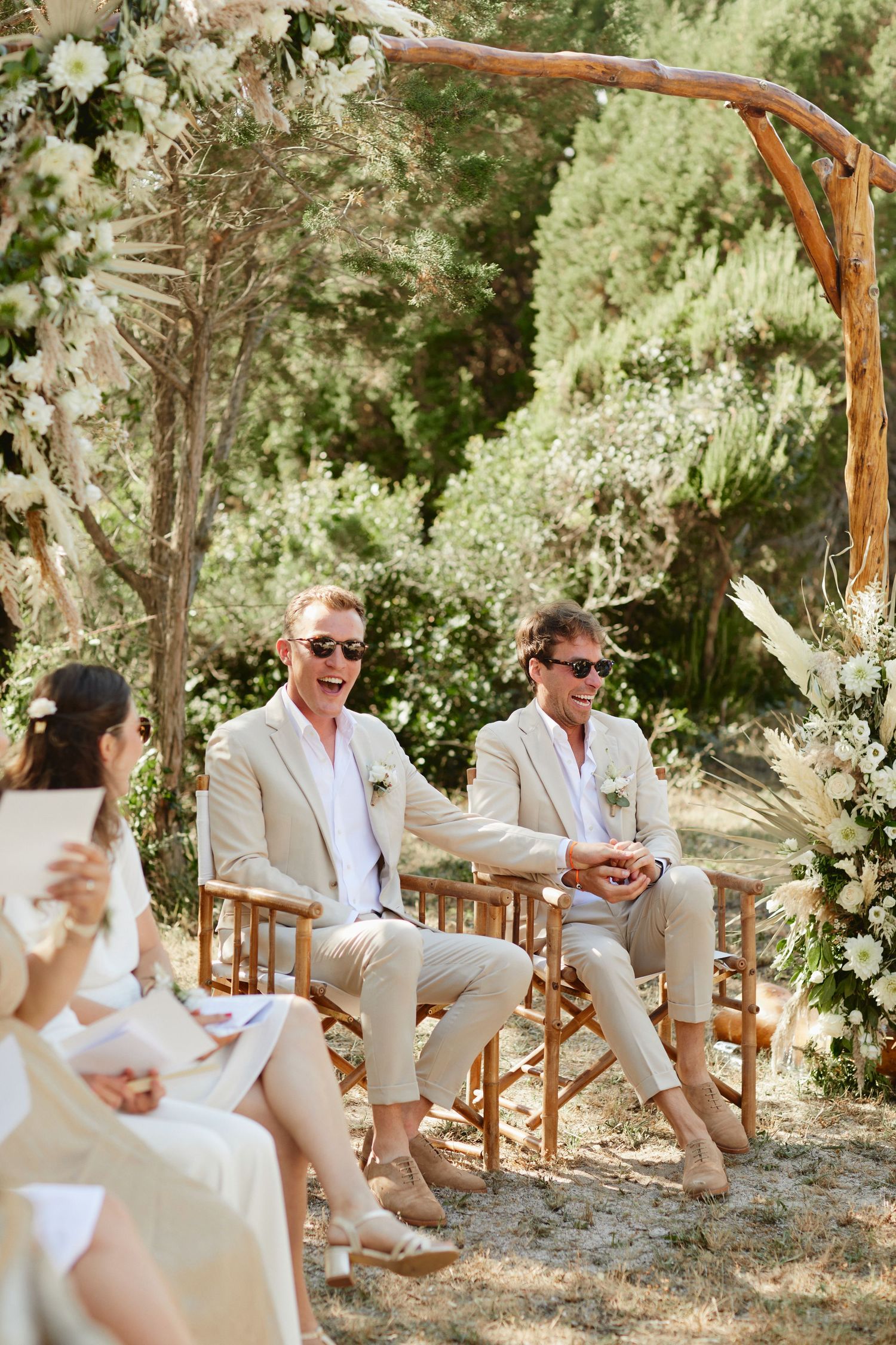 Two grooms in beige suits smile and hold hands under a floral arch at their outdoor wedding.