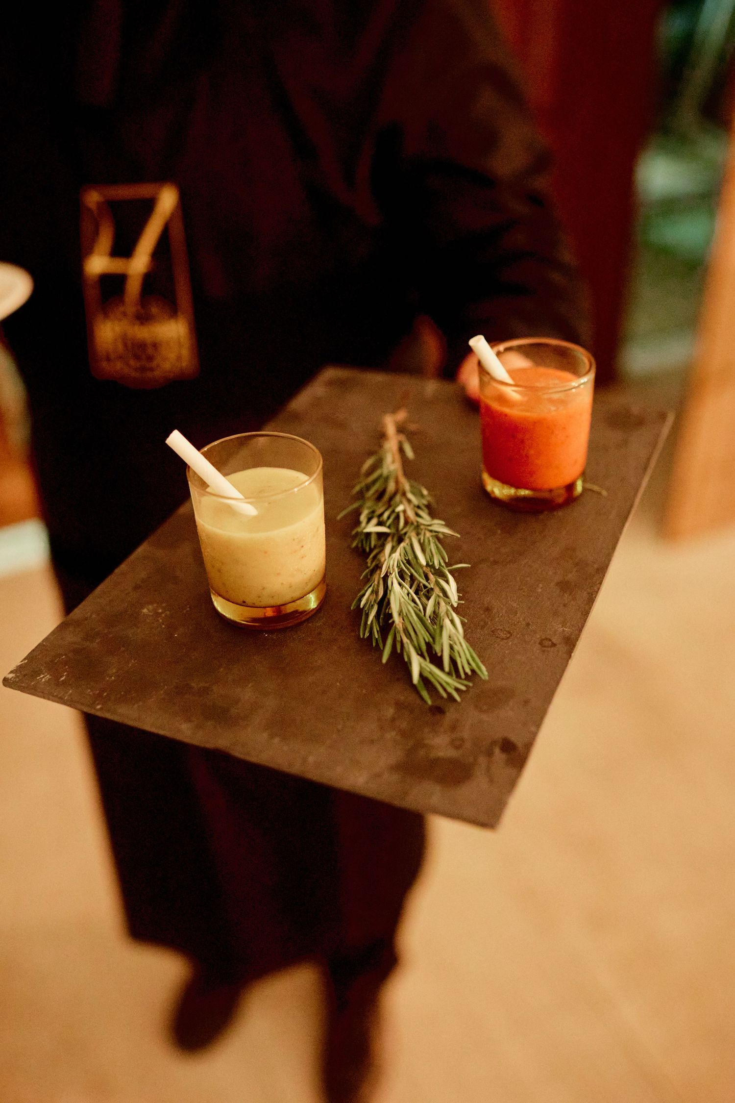 Waiter serving two cocktails on a dark tray with rosemary garnish, one yellow and one red drink with paper straws.