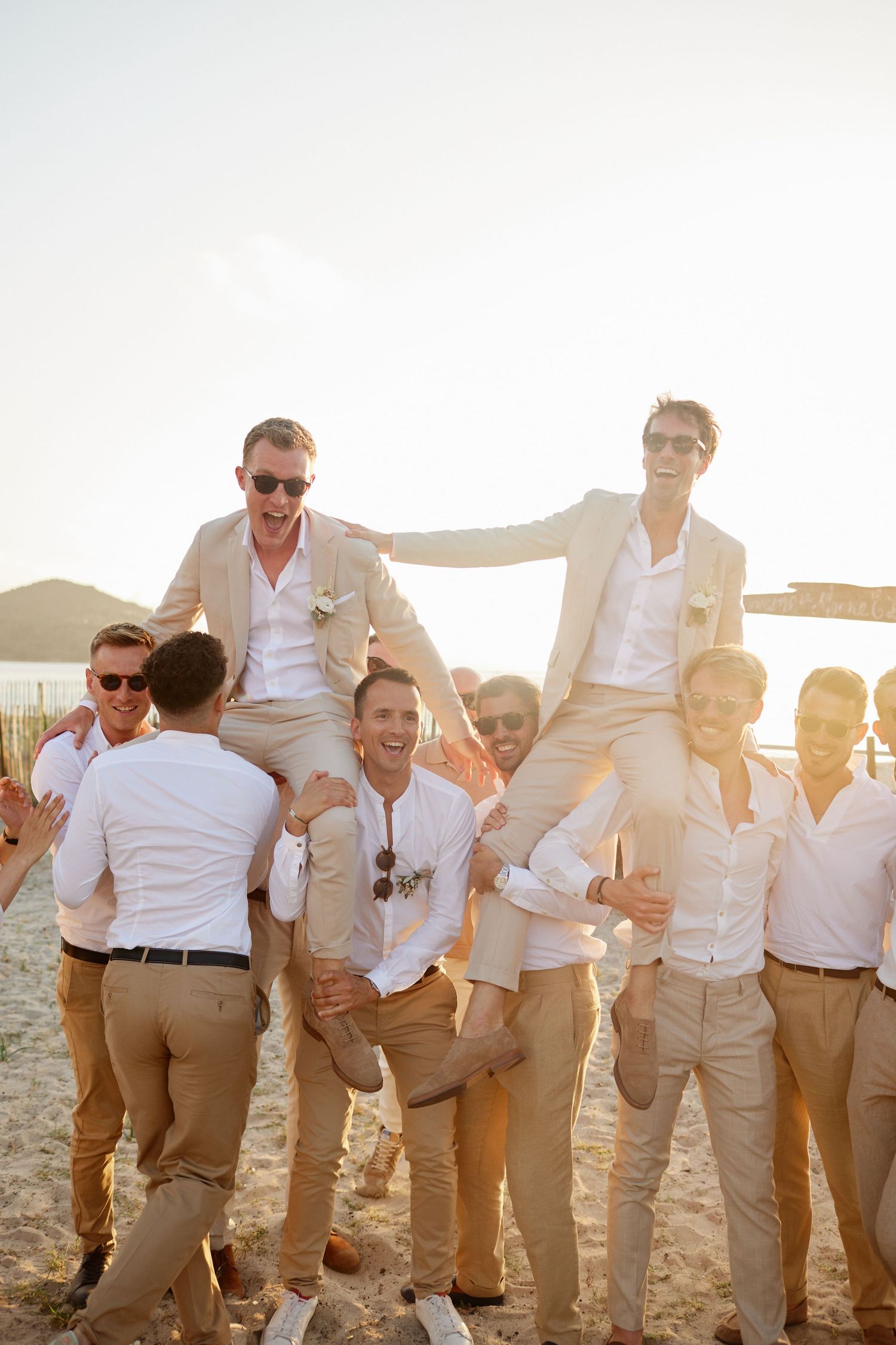 Group of groomsmen celebrating on a beach at sunset, two being lifted on shoulders in tan suits.