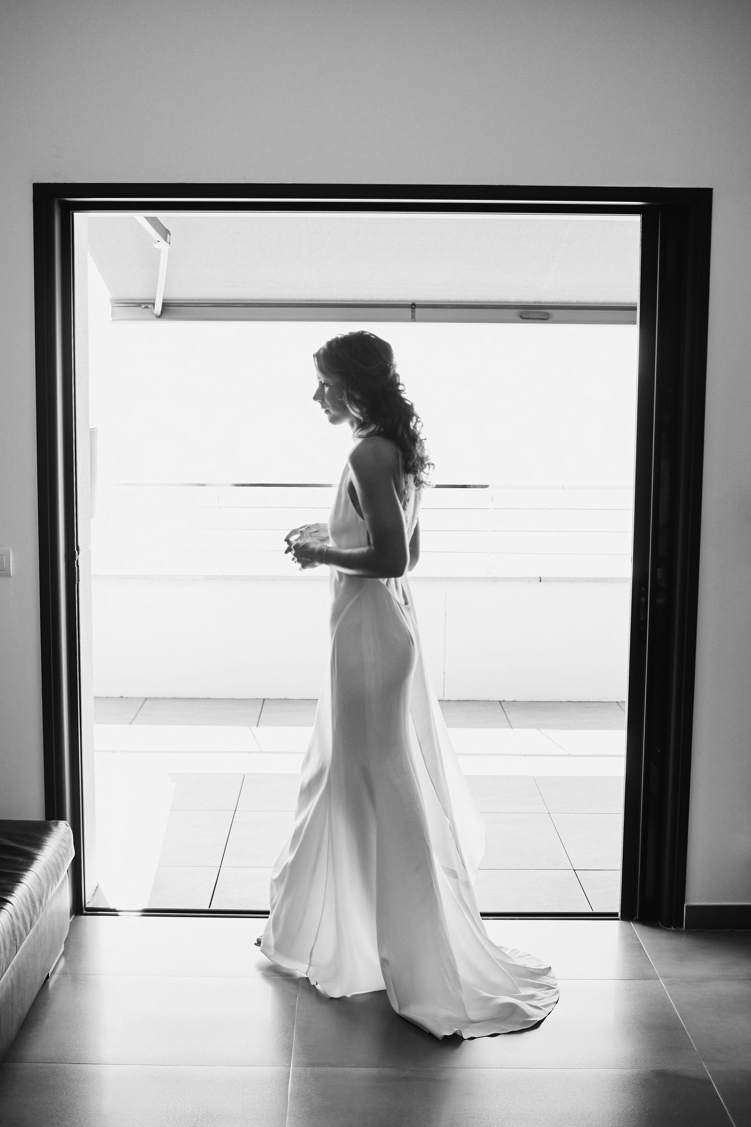 Elegant black and white silhouette of bride in fitted wedding gown standing in doorway with bright backlight.