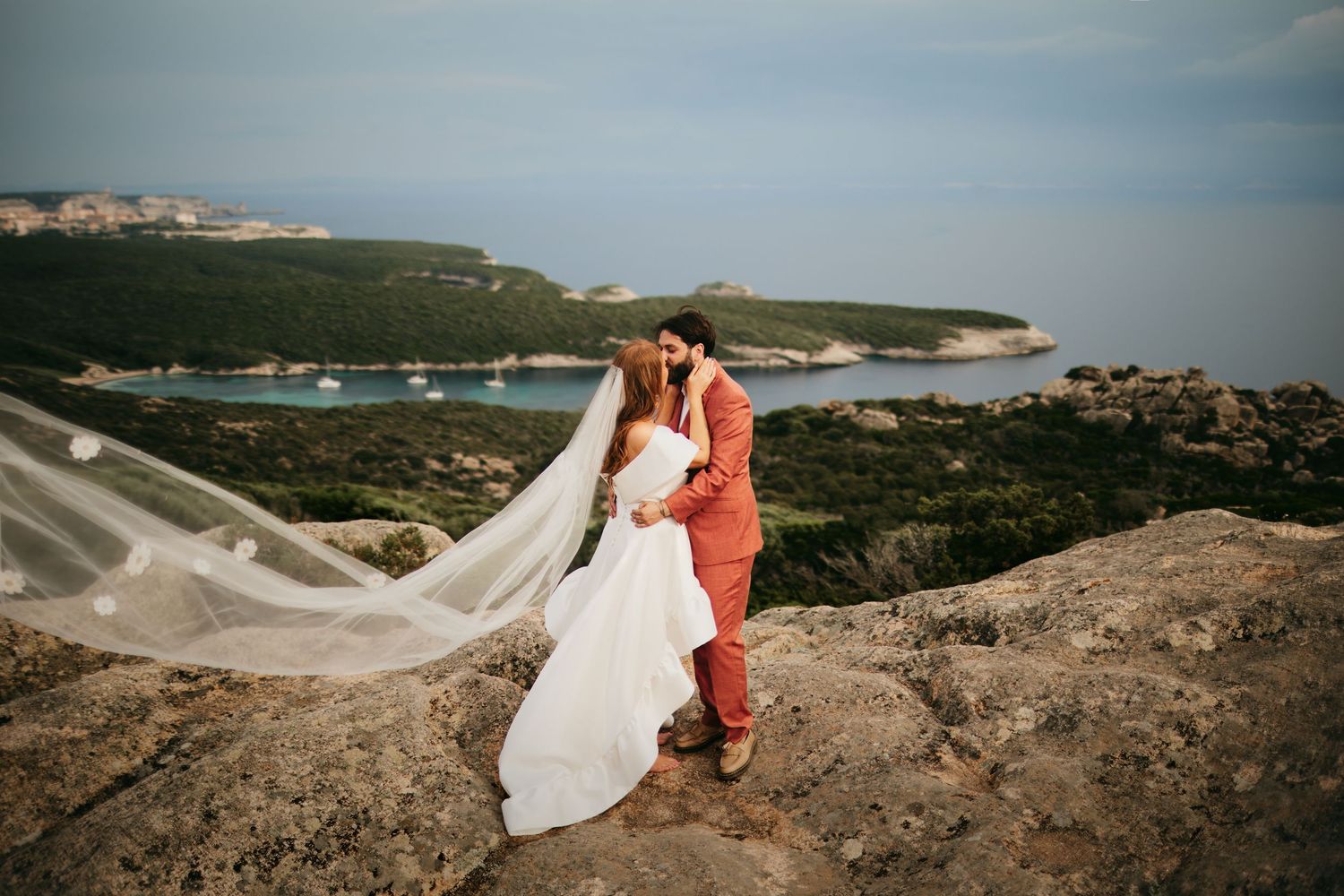 Bride and groom kissing on rocky hilltop with dramatic veil blowing in wind and coastal panorama below.