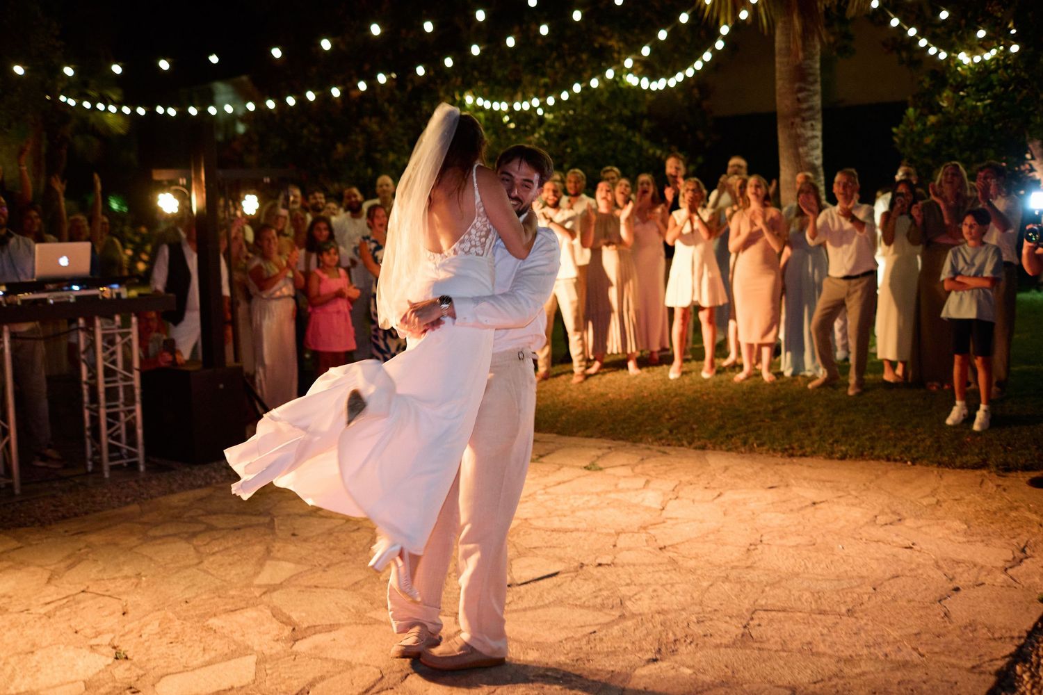 Groom lifts bride in white gown during first dance at outdoor wedding reception with cheering guests.