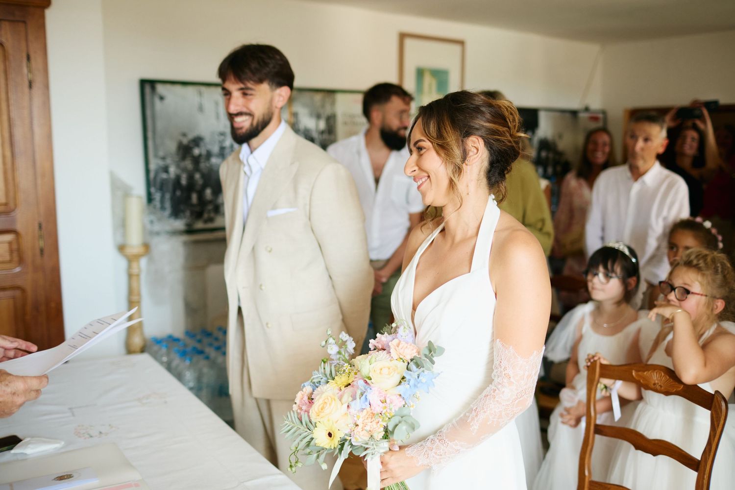 Smiling bride holding pastel bouquet stands beside groom in beige suit during indoor wedding ceremony.