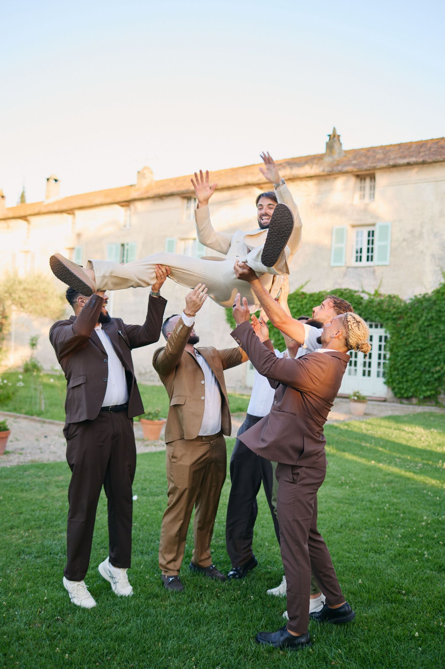 Groomsmen tossing laughing groom into the air on lawn outside stone chateau.