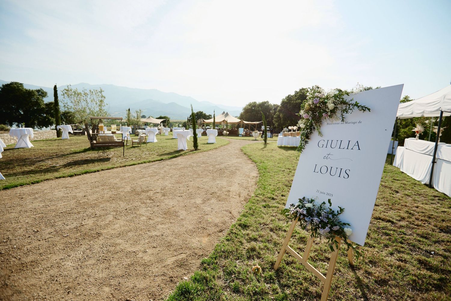 Outdoor wedding reception sign reading Giulia and Louis with mountain backdrop and white tents.