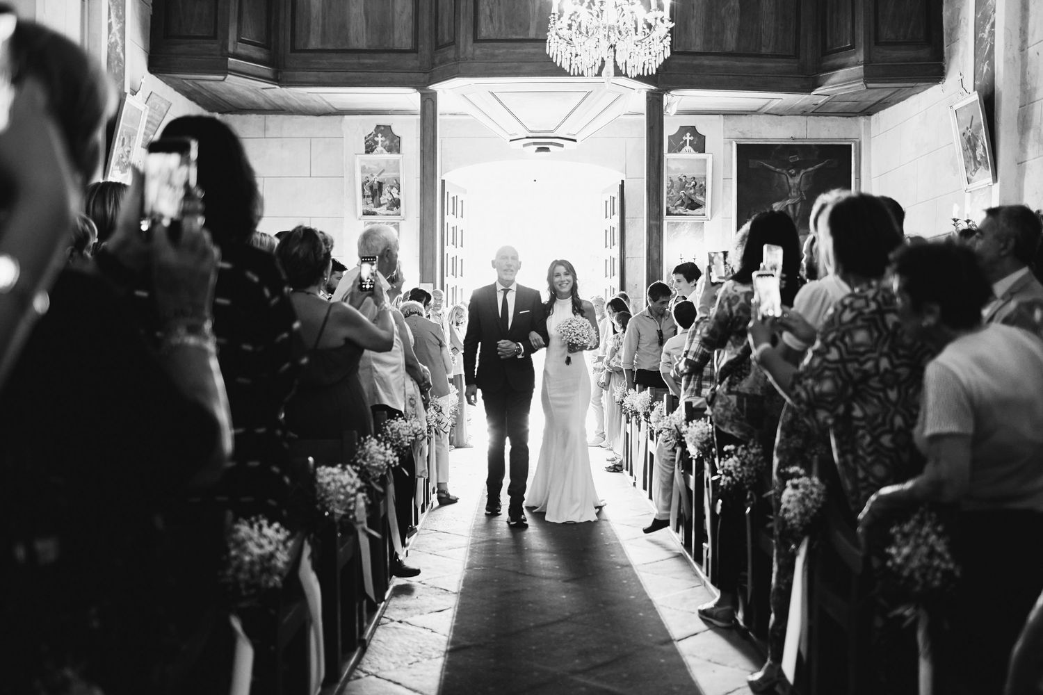 Black and white wide shot of bride walking down church aisle with escort through rows of seated wedding guests.
