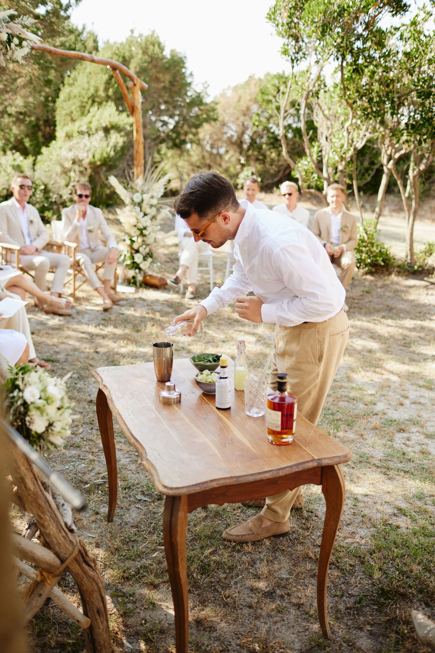 Man in white shirt prepares cocktails at wooden table during outdoor wedding ceremony in nature.