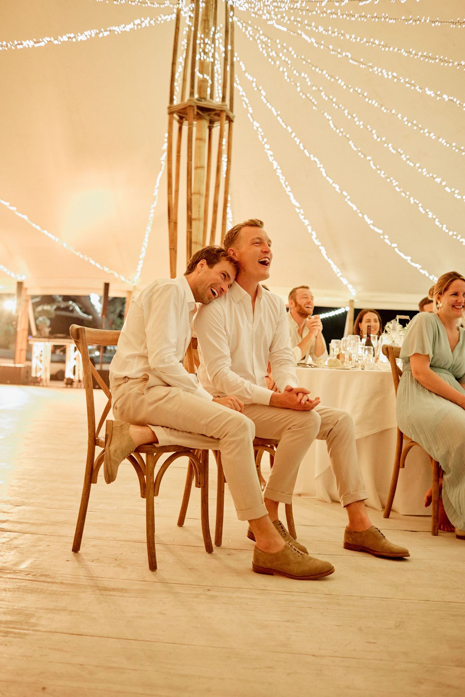 Two grooms sharing a tender moment laughing together at their wedding reception under twinkling fairy lights.