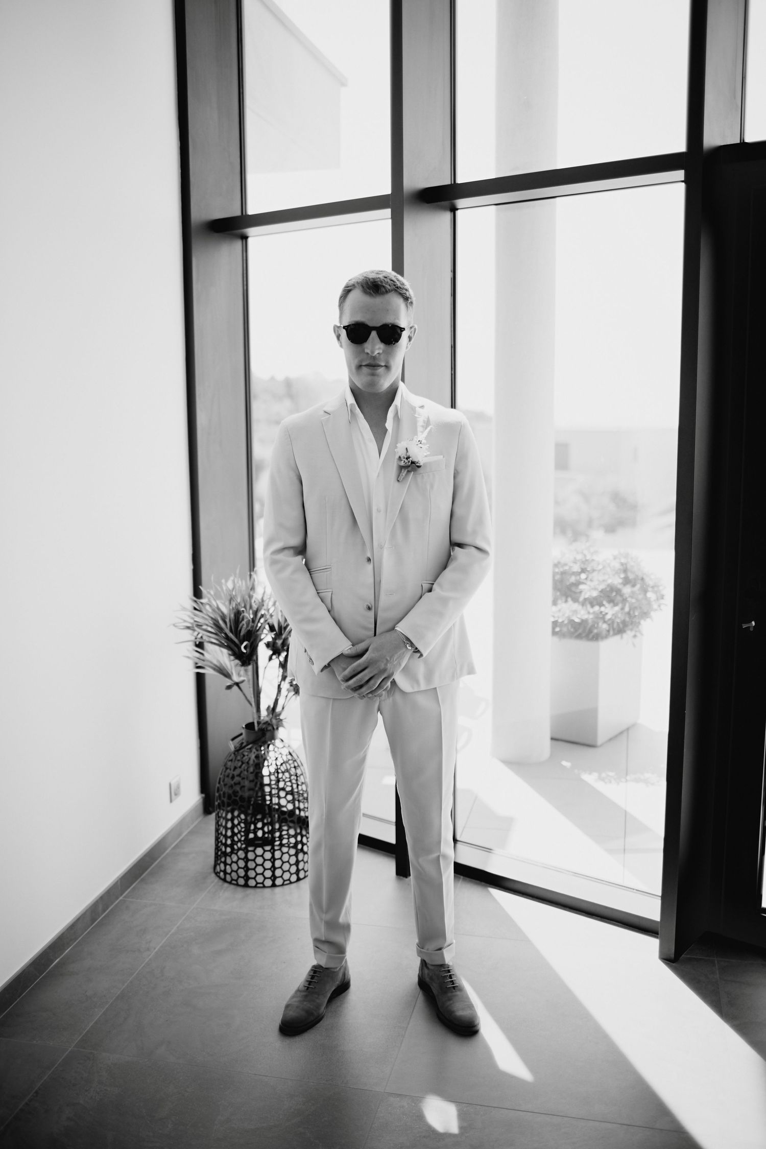 Groom in beige linen suit and sunglasses standing confidently in black and white full-length portrait.