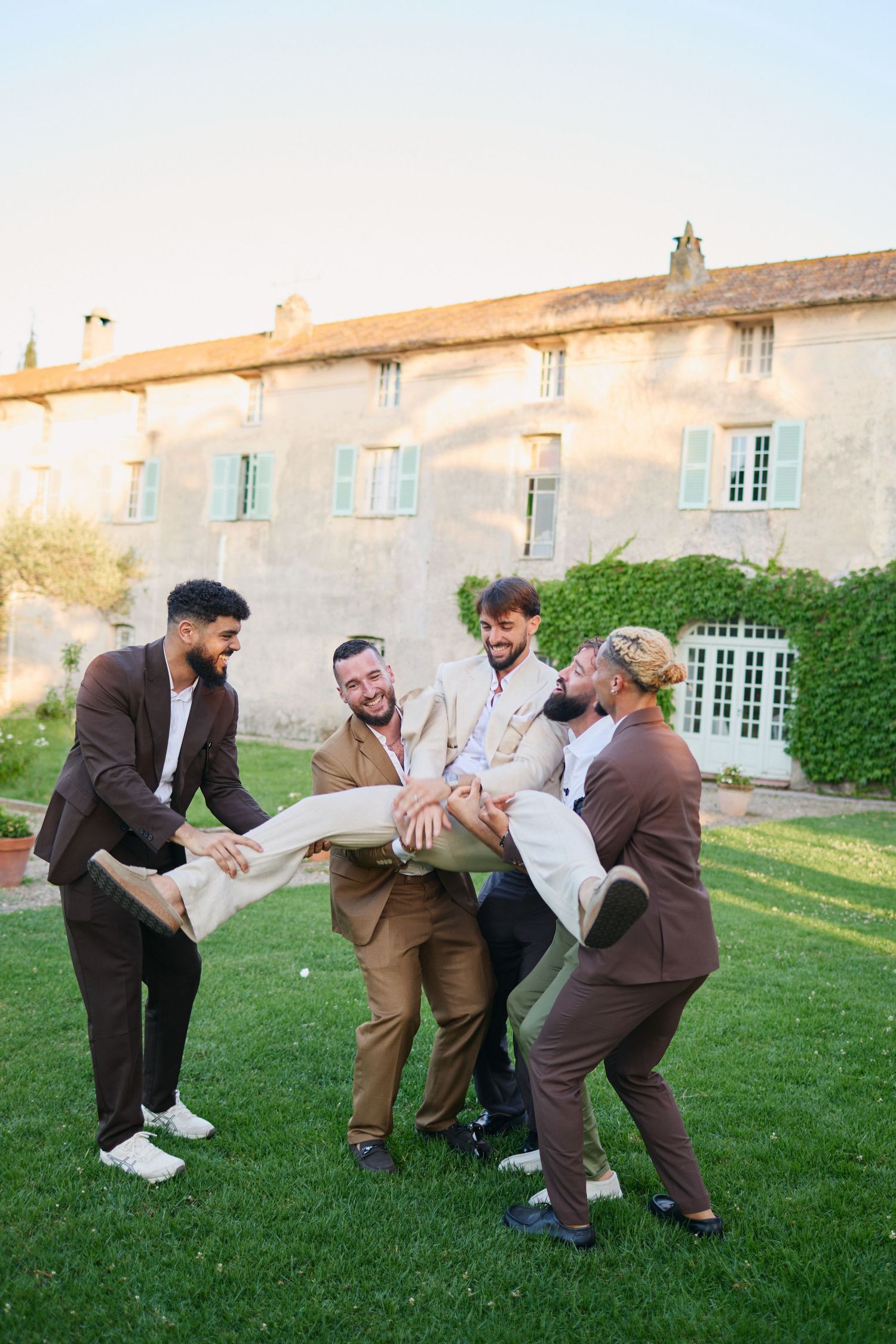 Laughing groomsmen carrying groom horizontally on grass outside stone French chateau.