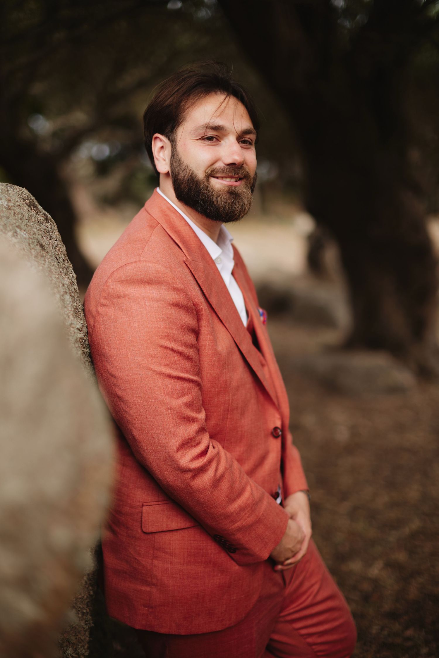 Handsome bearded groom in a stylish terracotta linen suit posing outdoors among ancient trees.