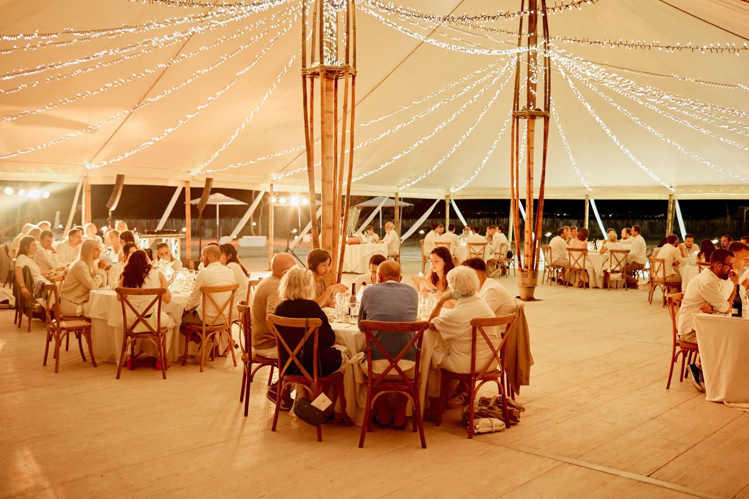Elegant outdoor wedding reception with guests seated at round tables under a warmly lit sailcloth tent.