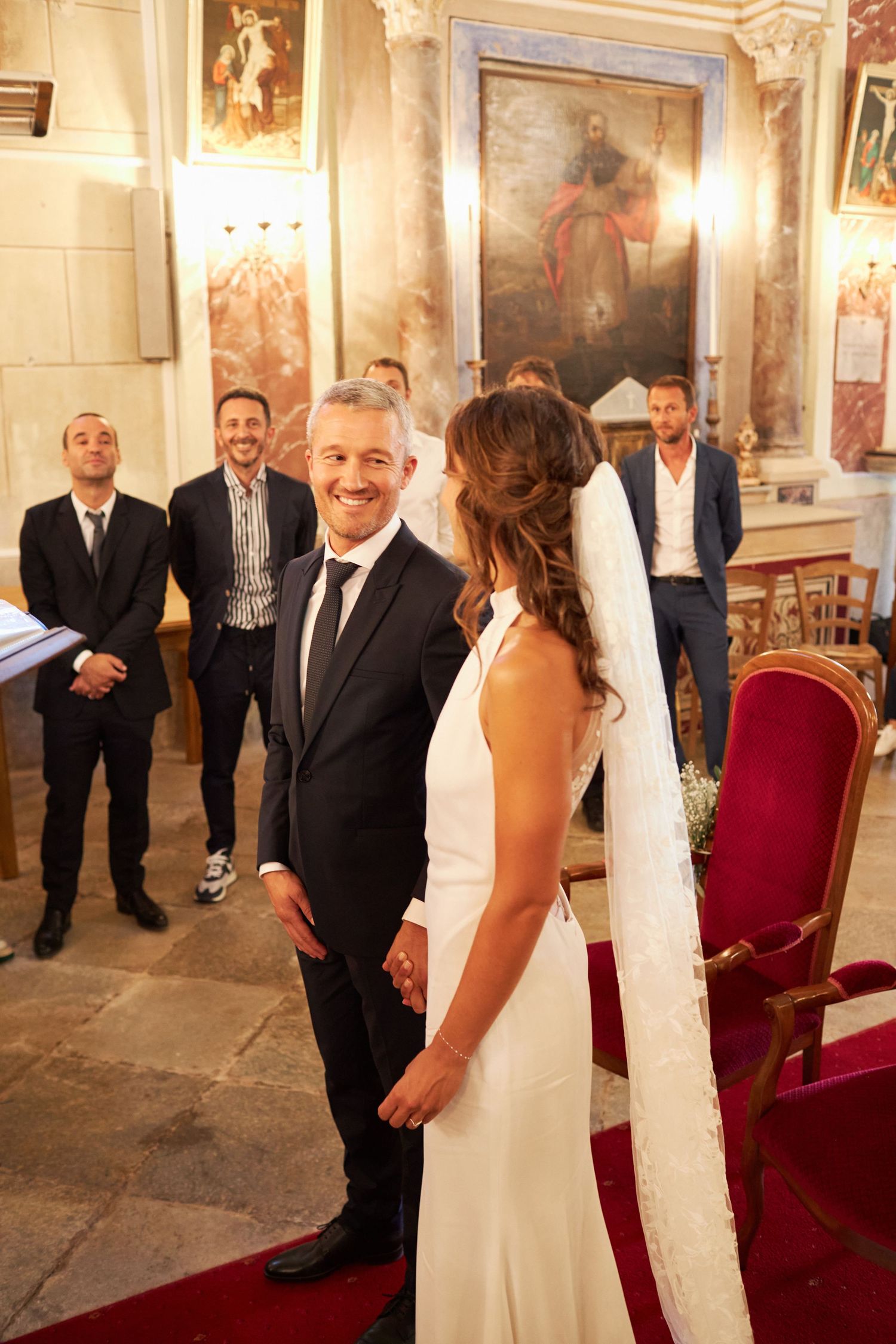 Groom in black suit smiles at bride in white dress at church altar with guests behind.