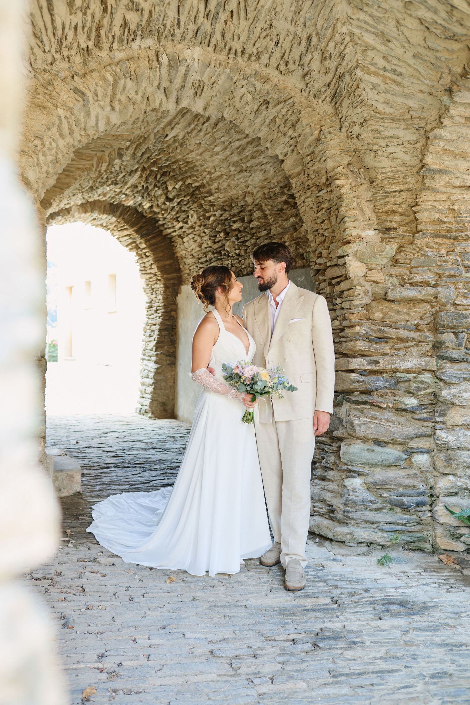 Bride and groom share romantic moment under ancient stone archway with bouquet.