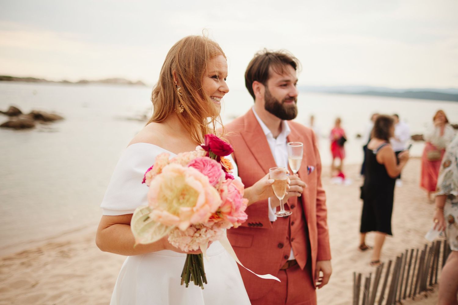 Bride holding pink bouquet and groom in coral suit walking on beach during sunset wedding ceremony.