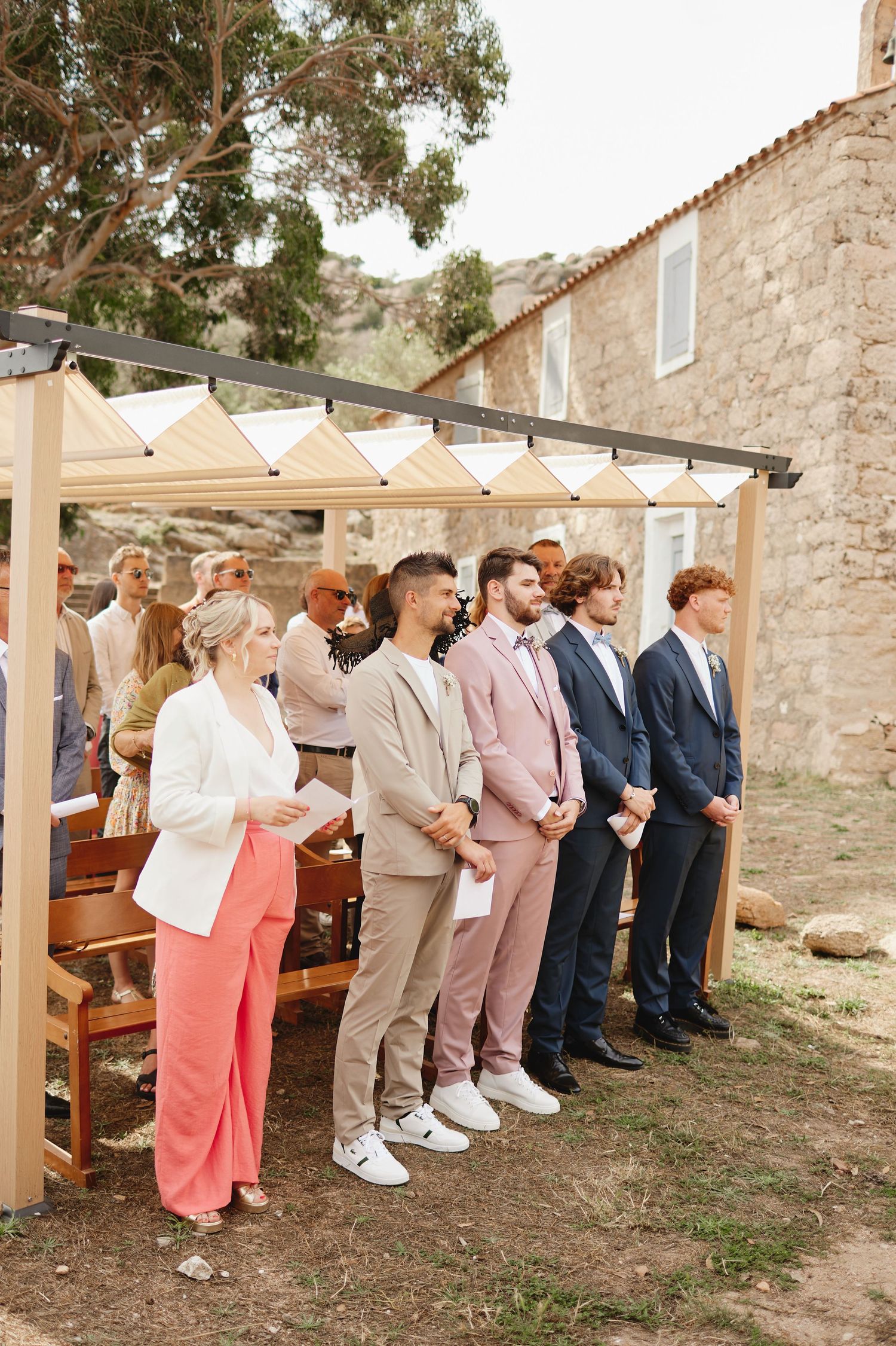 Wedding party stands in line during outdoor ceremony beside stone building on sunny day.