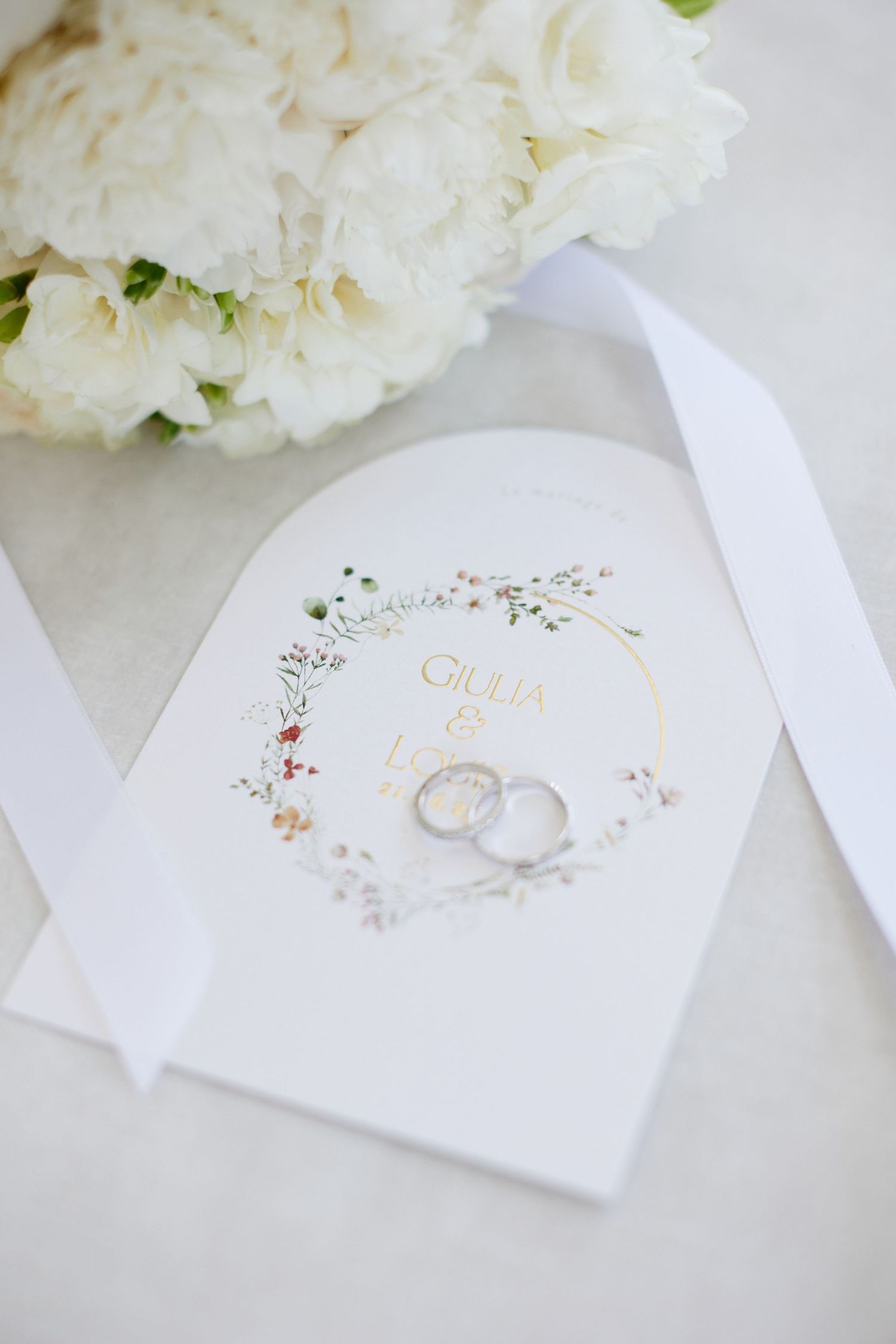 Wedding rings resting on ceremony booklet reading Giulia and Lorenzo beside white peony bouquet.