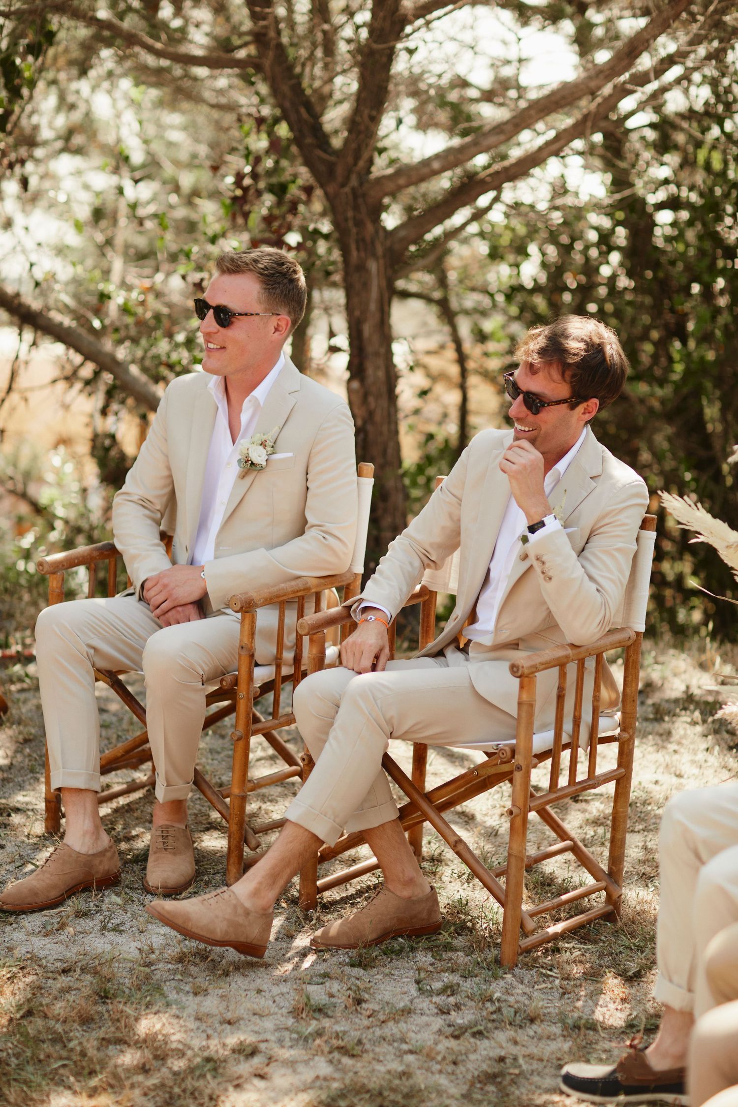 Two grooms in tan suits and sunglasses seated at outdoor wedding ceremony.