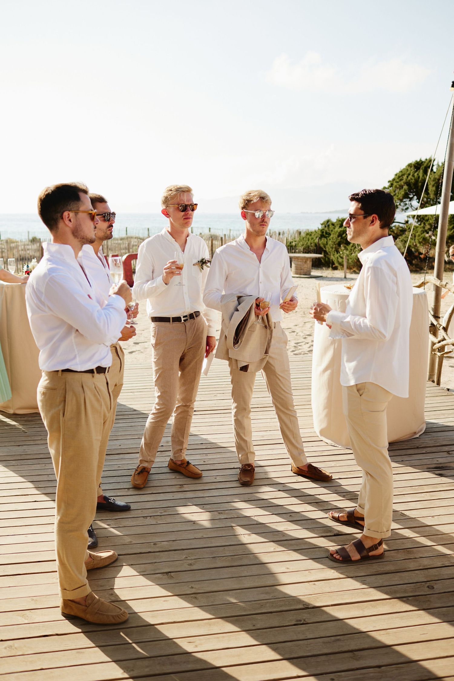 Groomsmen in white shirts and khaki pants socializing on a wooden deck at a beach wedding.