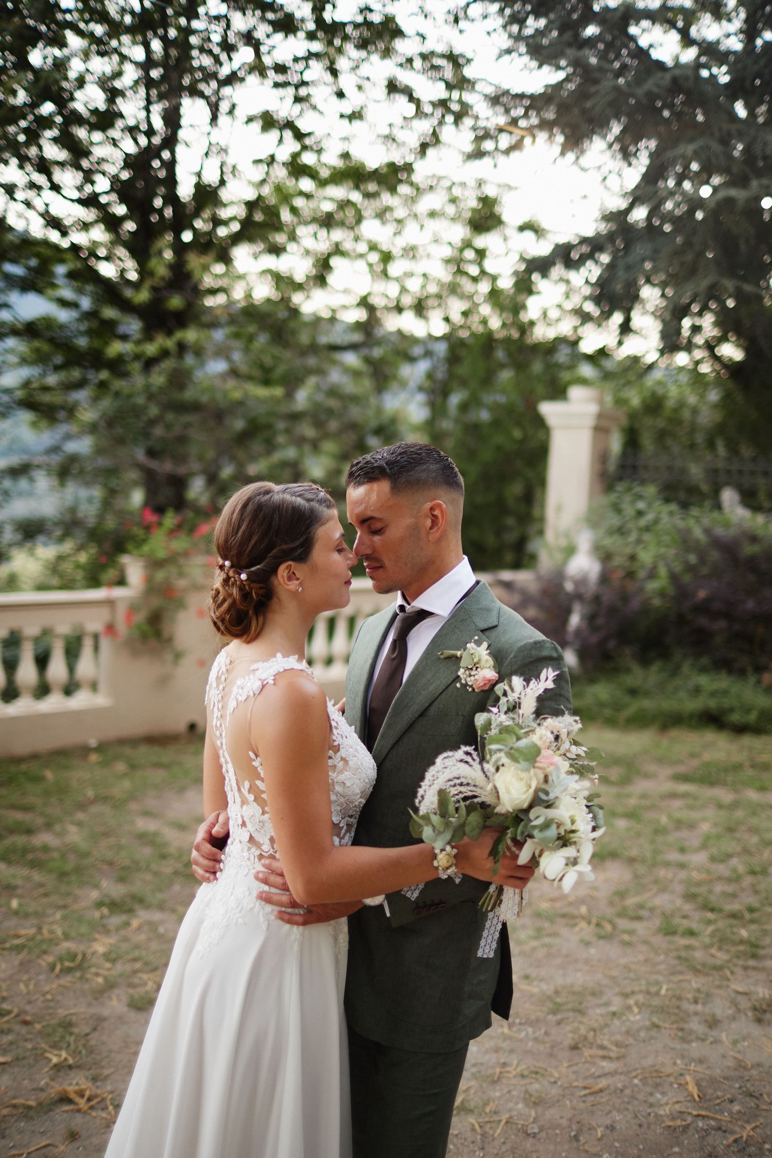 Bride and groom share an intimate moment in a lush garden, she in lace gown, he in olive suit holding bouquet.