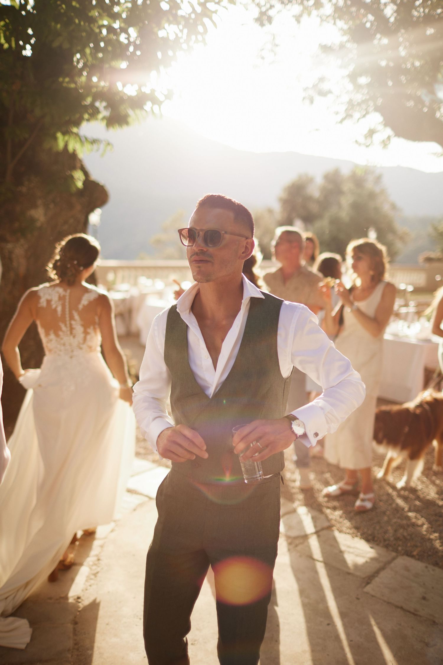 Stylish man in vest and sunglasses dancing at outdoor wedding reception in golden light.