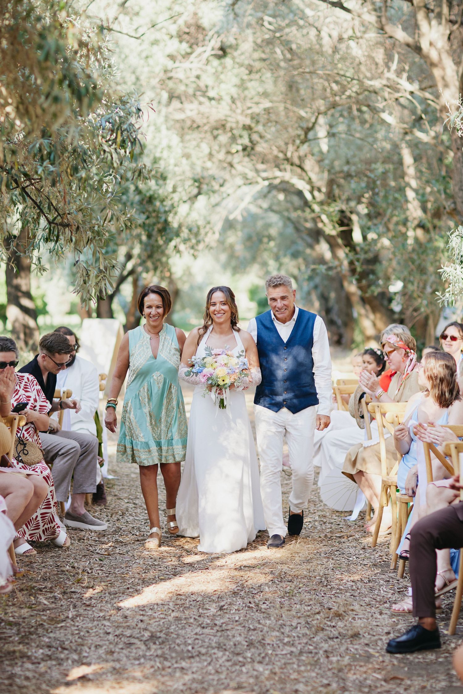 Smiling bride in white gown walks aisle with bouquet escorted by parents at outdoor wedding.