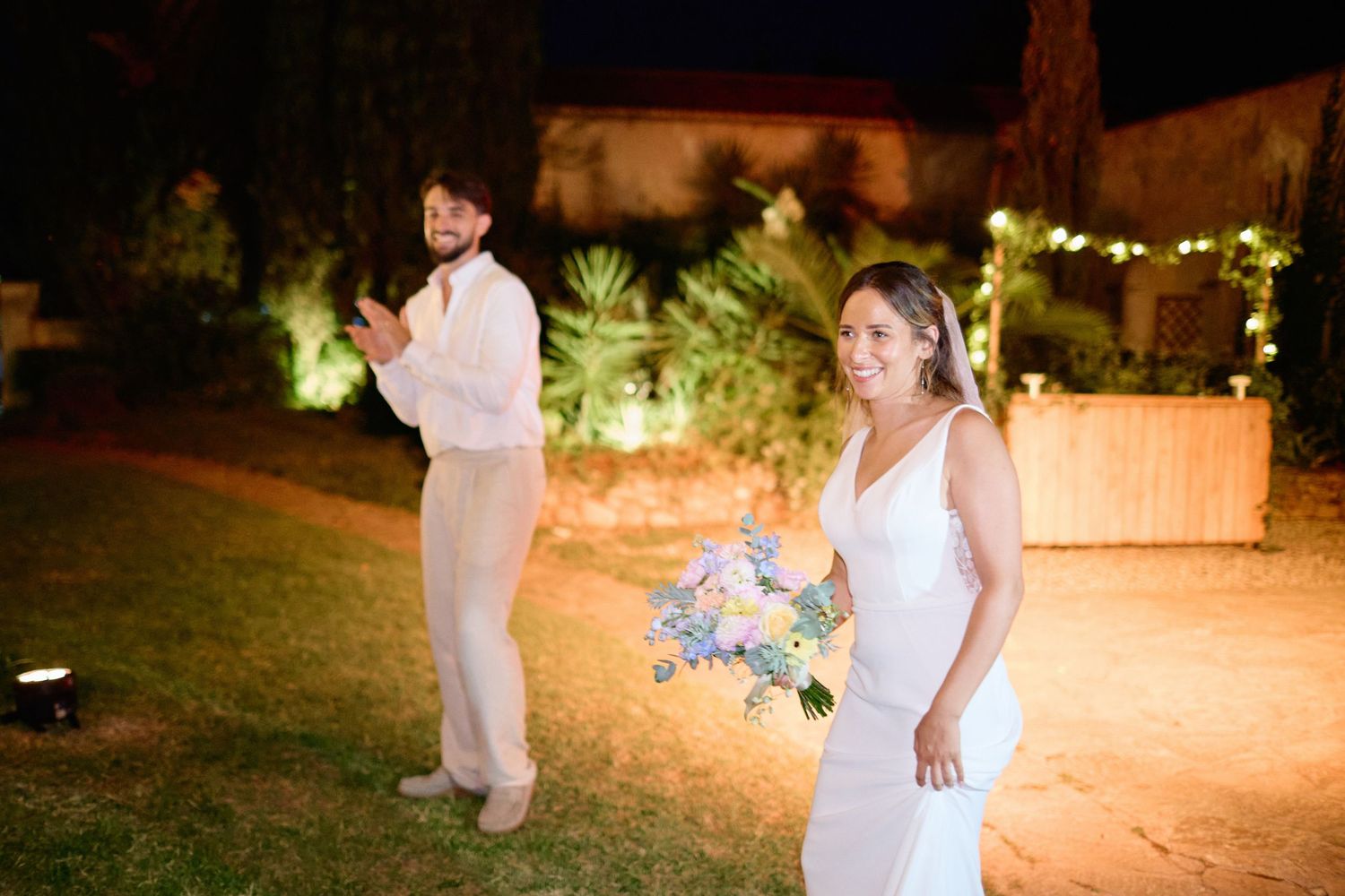 Bride holding colorful bouquet standing on lawn at night with groom in background.