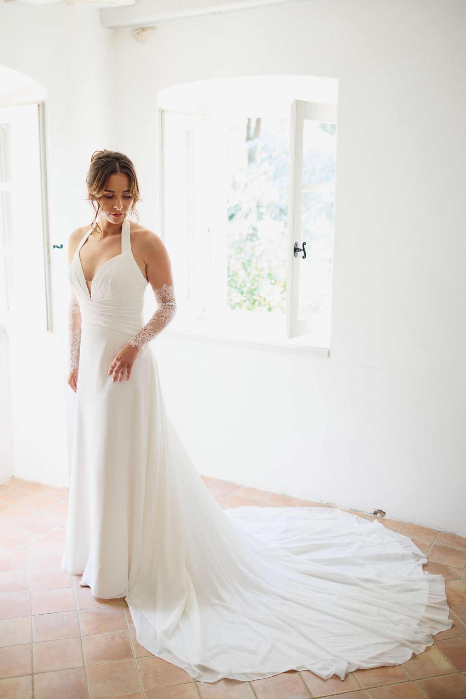 Bride in white halter wedding dress with long train standing gracefully in sunlit white room.