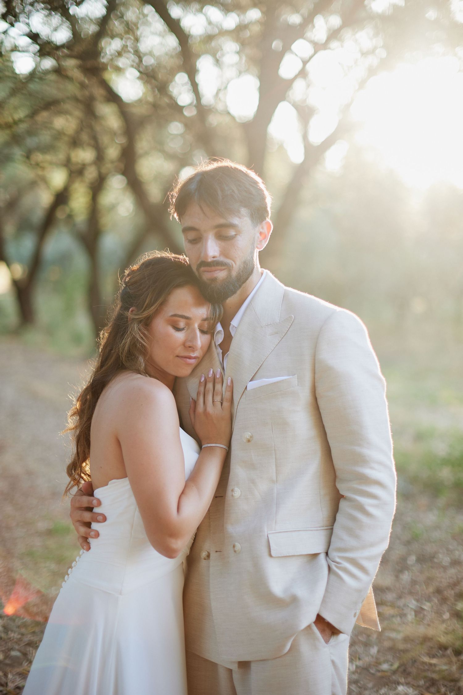 Bride resting her head on grooms chest in soft backlit sunlight among trees in a romantic outdoor setting.