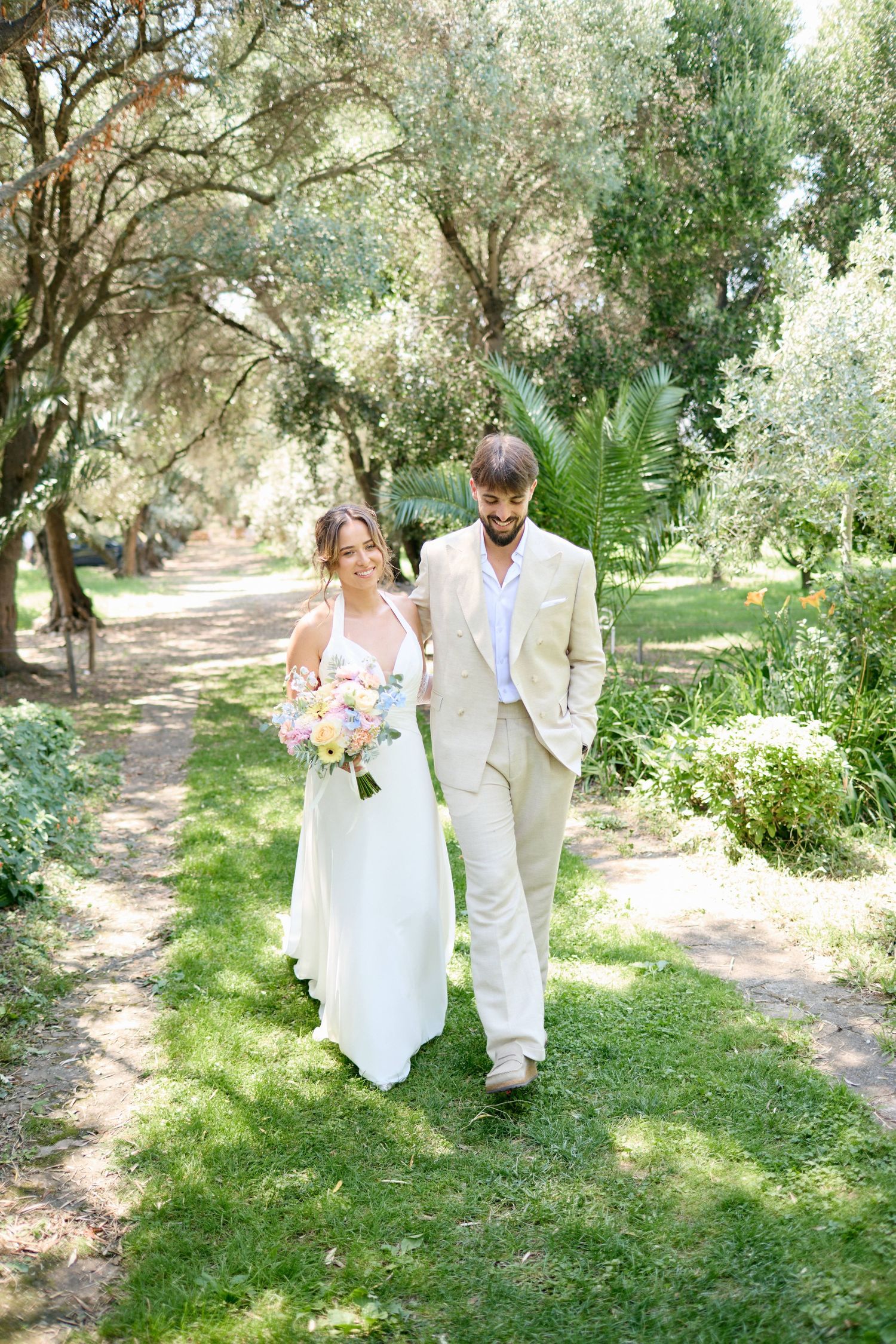 Bride and groom walking hand in hand through a lush green tree-lined garden path.