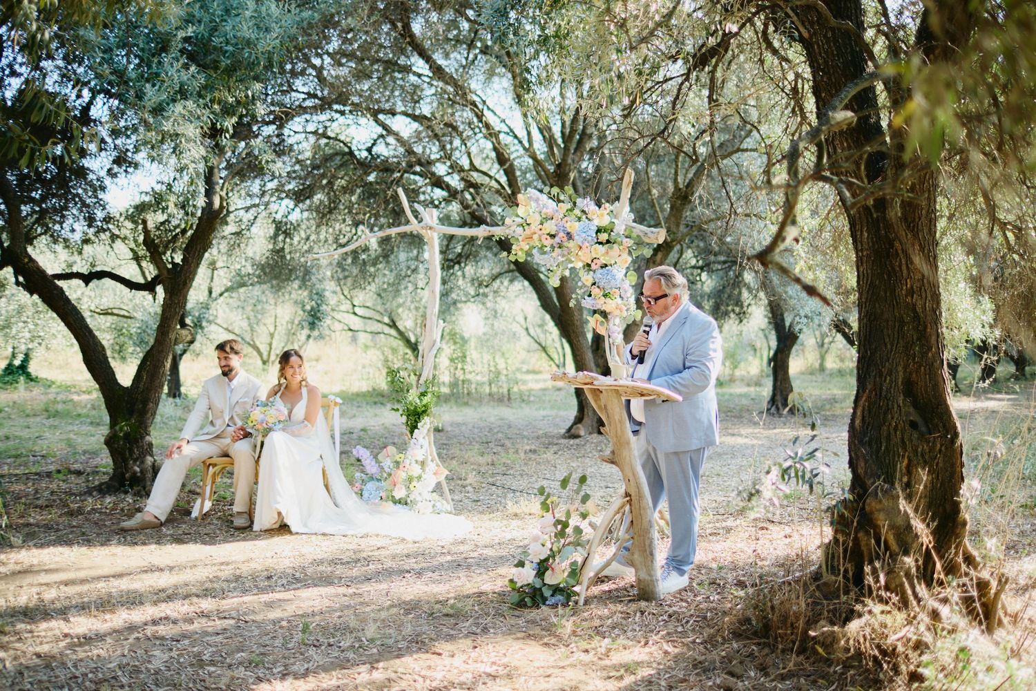 Officiant reads at wooden podium as bride and groom sit at rustic outdoor wedding ceremony.
