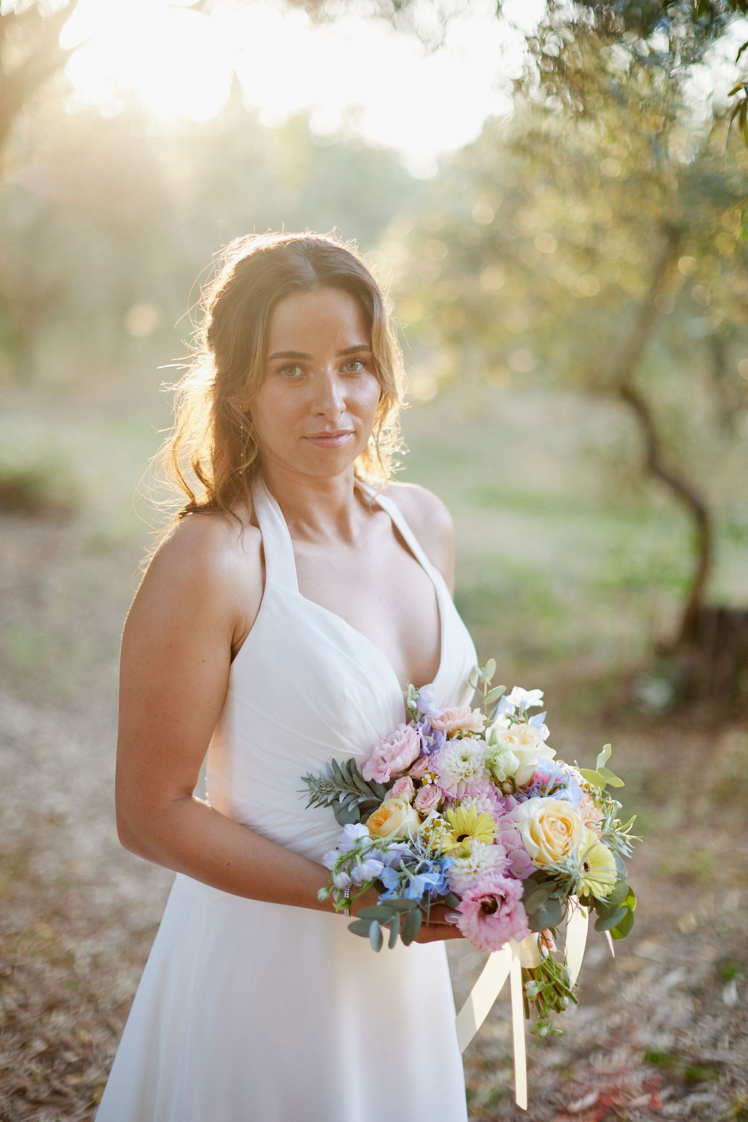 Bride in deep V-neck white halter gown holding a pastel bouquet, posing in a sun-drenched olive grove.