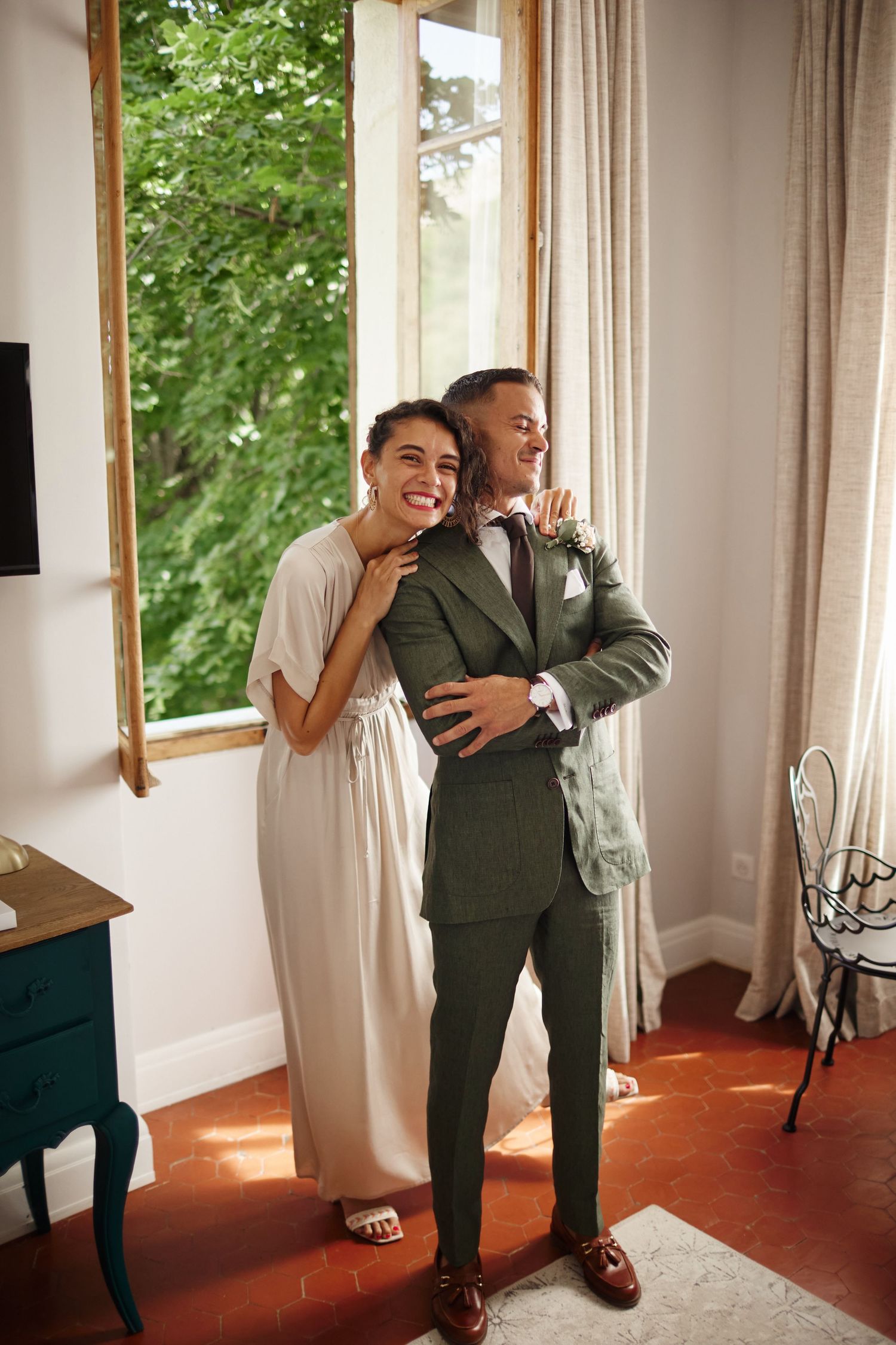 Smiling bride in ivory dress embraces groom in green suit by sunlit window indoors.