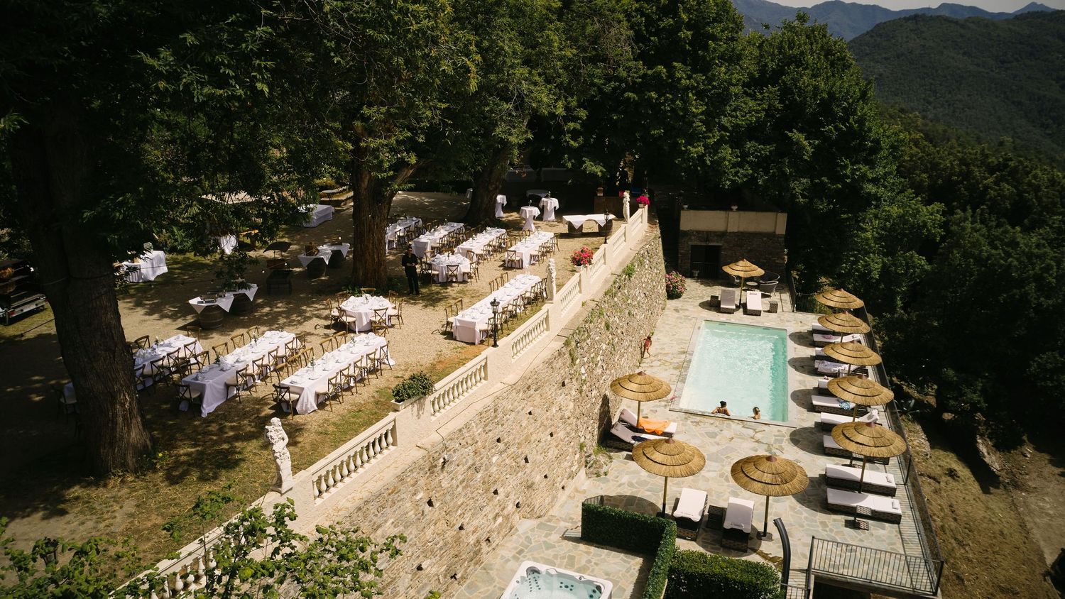 Aerial view of an elegant outdoor wedding reception with white tables beside a pool at a stone villa.